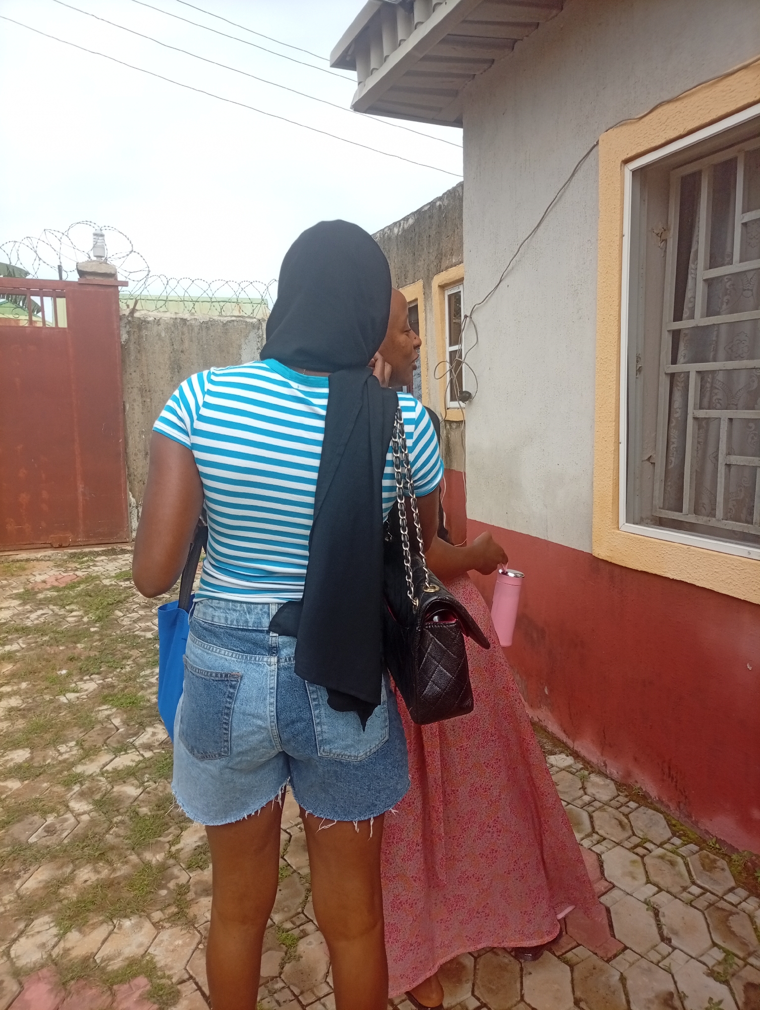 In the city of Unknown, Unknown, two women are standing outside a building. The woman on the left is wearing a blue and white striped t-shirt, denim shorts, and a black hijab. She is carrying a blue bag and a black purse. The woman on the right is wearing a pink patterned dress and is holding a pink insulated cup. The building appears to be a house with a red and tan facade and a window with metal bars. A barbed wire fence is visible in the background. The ground is paved with interlocking concrete blocks. The sky is overcast.