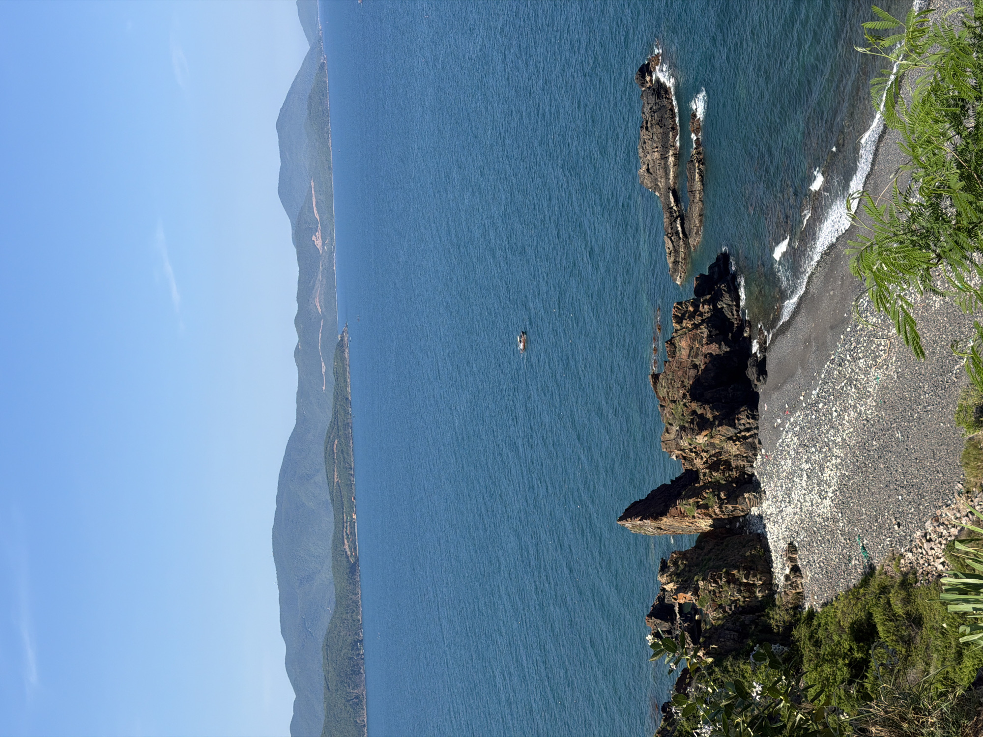 This is a long shot, eye-level, daytime, outdoor photograph taken in Nam Nha Trang Ward, Vietnam. The image features a vast expanse of deep blue ocean under a clear, bright blue sky. In the foreground, a rocky coastline with some vegetation leads down to a pebble beach. The beach is covered in a mix of dark and light-colored pebbles, with a notable amount of scattered debris, including what appears to be plastic waste. Jagged, dark brown rock formations jut out of the water near the shore, with some waves lapping against them. In the mid-ground, a small, orange and white boat is visible on the water. In the background, a series of lush, green, mountainous islands stretch across the horizon, with the hazy outline of more distant landmasses further beyond. The overall scene depicts a coastal landscape with a significant environmental concern due to the pollution on the beach.