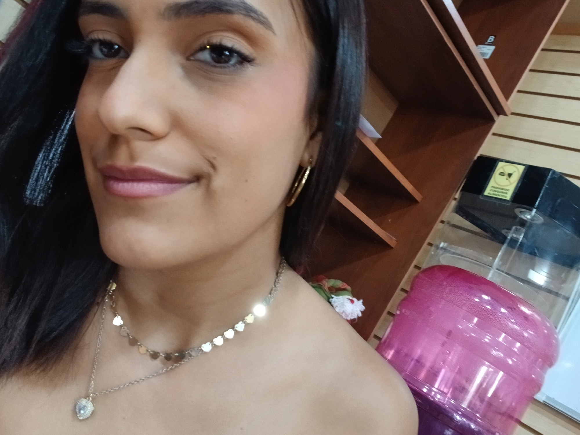 A close-up selfie shot of a woman with dark hair, light skin, and brown eyes. She is wearing gold hoop earrings and two delicate necklaces, one with small heart-shaped pendants and the other with a heart-shaped pendant. The background includes a wooden shelf with various items, including what appears to be a water dispenser with a pink container. The location, Juan Griego, Venezuela, is not evident in the image itself and would require additional contextual information.