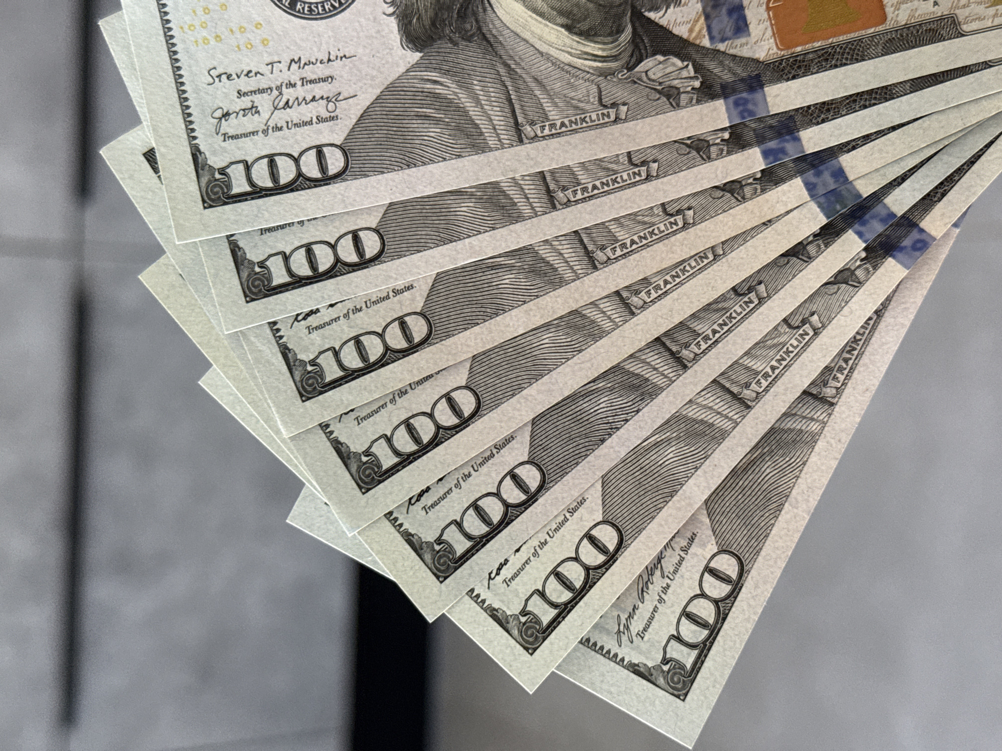 The image shows a close-up, fan-like arrangement of several US $100 bills. The bills are overlapping and angled, with only portions of each visible. The bills appear to be crisp and new, with clear printing of the denomination "100" and the portrait of Benjamin Franklin. The background is a blurry, neutral gray, providing no specific environmental context. There is no indication of any connection to the city of Lyubertsy, Russia.