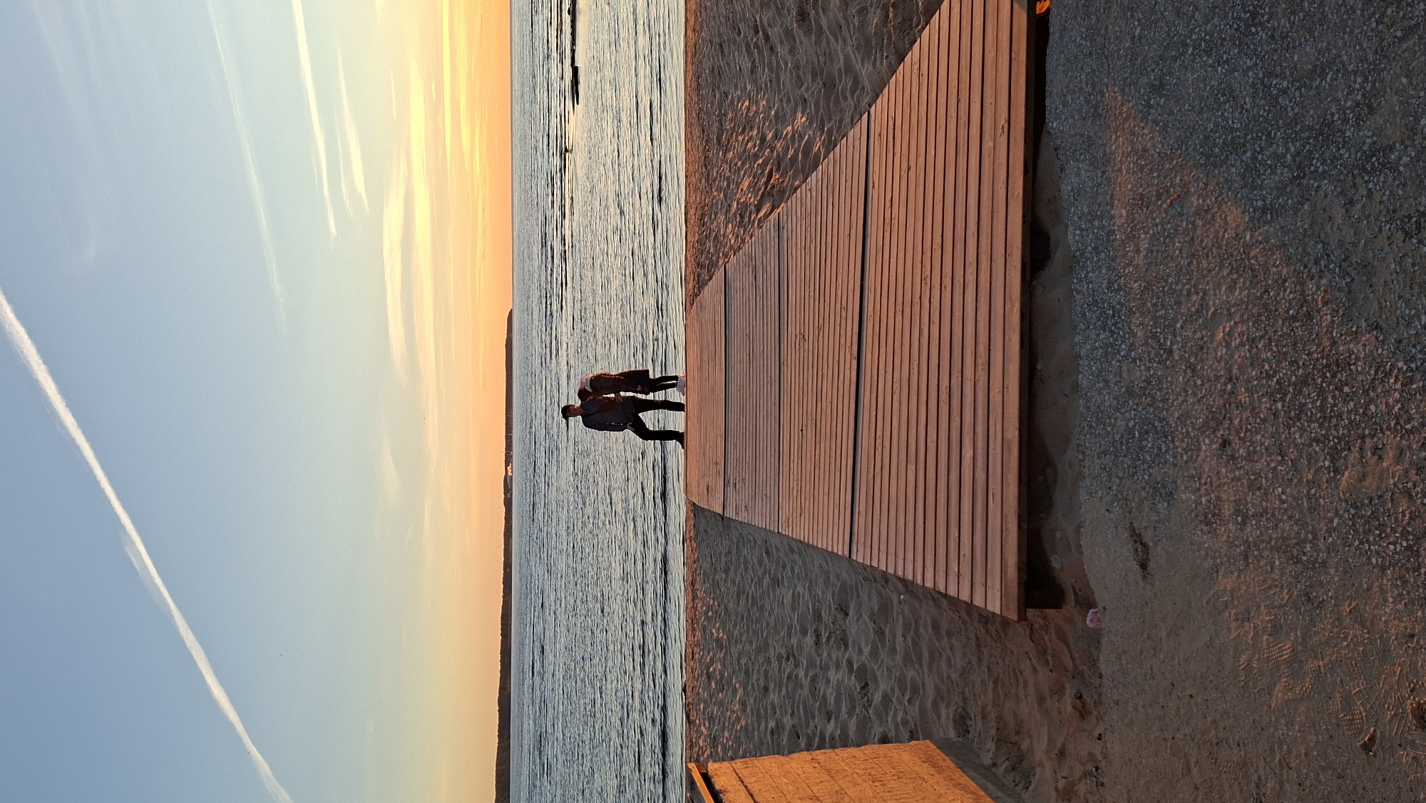 At sunset on a beach in Tallinn, Estonia, a man wearing a cap and dark clothing stands to the left of a woman dressed in a brown coat. They are positioned on a wooden boardwalk that extends onto the sand. The boardwalk leads towards the sea, which is calm with gentle waves. In the distance, a landmass is visible on the horizon. The sky is a gradient of blue and orange, with a long, thin contrail stretching across it. The sand is lightly textured, and shadows are cast by the boardwalk and a nearby structure on the left.