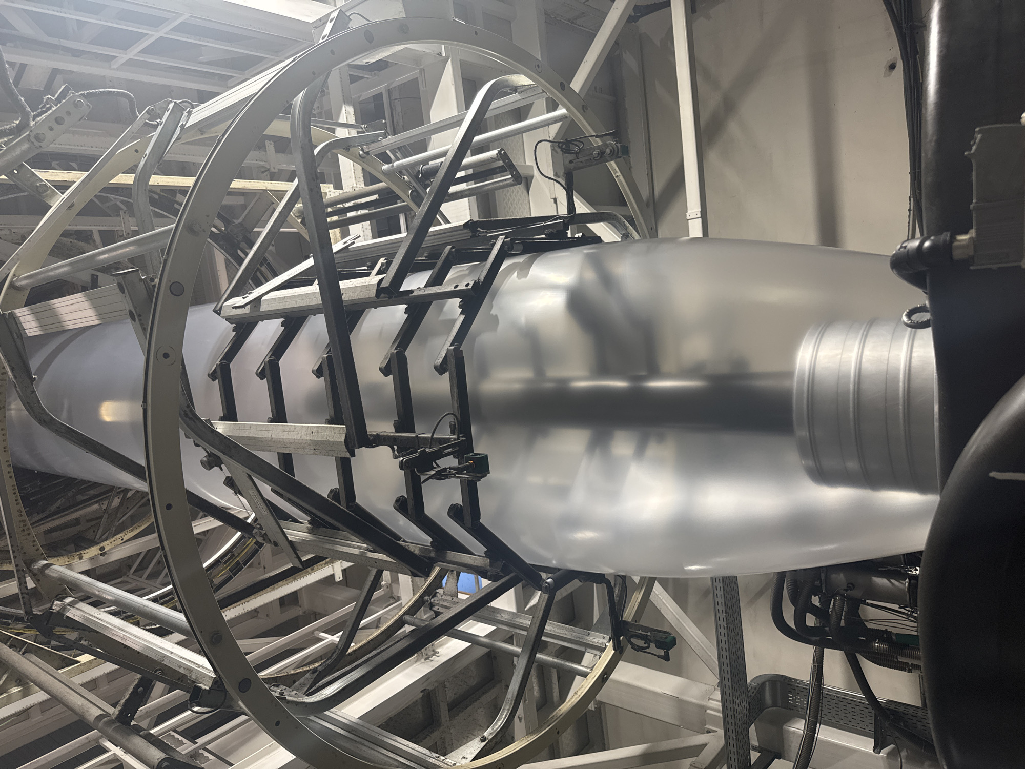 The image captures an industrial manufacturing process, likely within a factory setting in Perm, Russia. The central subject is a large, partially inflated, translucent plastic film, formed into a cylindrical "bubble." This indicates a blown film extrusion process, where a polymer is expanded into a thin-walled tube.
The plastic bubble is supported and guided by an intricate framework of metallic structures. These consist of curved arcs, straight beams, and various support components, predominantly in shades of light gray, white, and darker metallic finishes. Numerous small, black, adjustable mechanisms or sensors are attached to this framework, likely used to control the shape and tension of the expanding film.
In the background, further industrial infrastructure is visible, including grid-patterned ceilings and vertical support beams, suggesting a large-scale facility. On the right side of the frame, dark, corrugated pipes and conduits with fittings are present, hinting at connections to machinery or fluid transfer systems. The entire scene is brightly illuminated by artificial lighting. No people or discernible text are visible in the image.