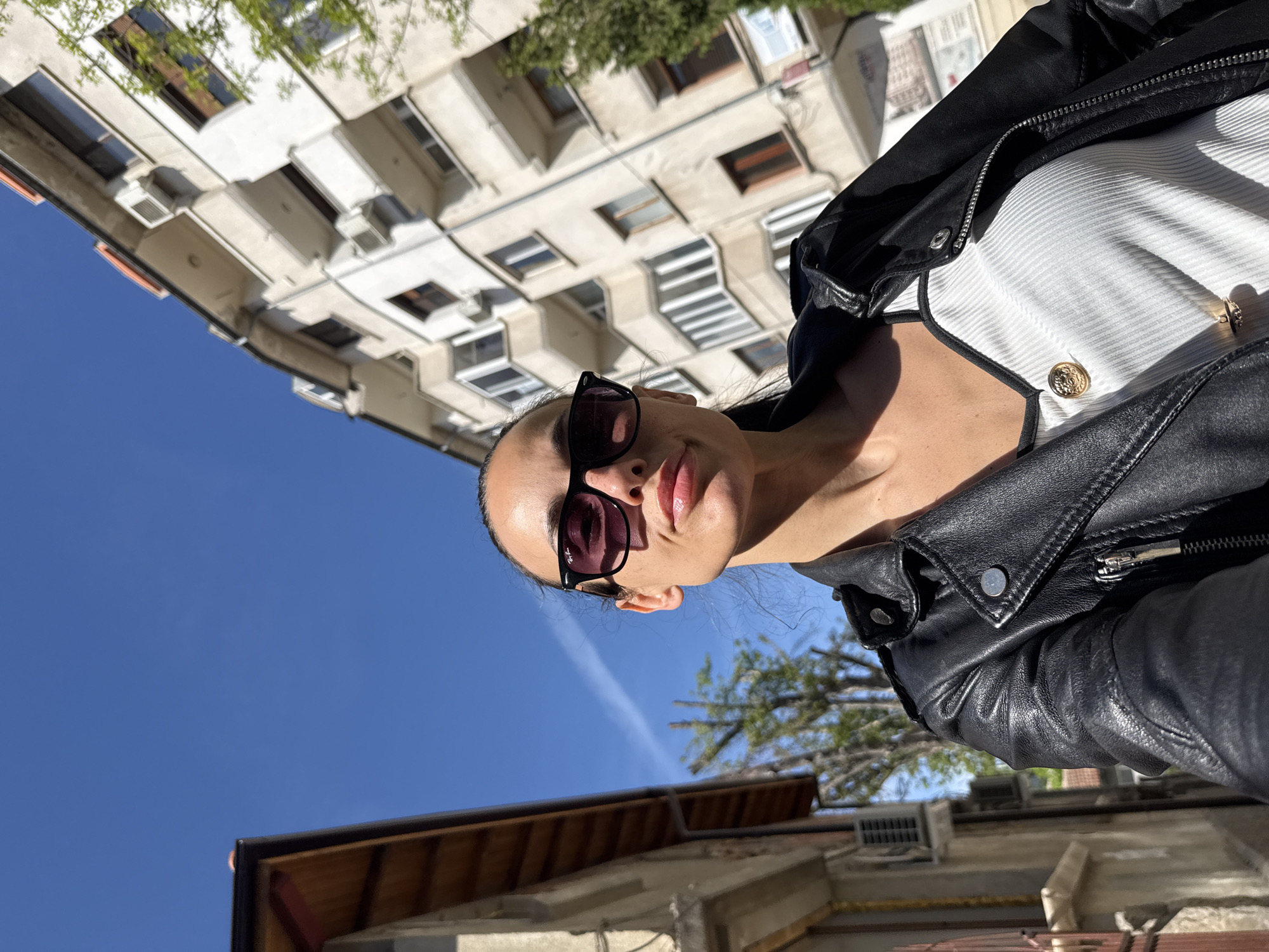 A low-angle selfie, rotated approximately 90 degrees clockwise, captures a person (likely female) from the chest up, looking upwards. The subject wears dark sunglasses with a visible "Ray-Ban" logo, a black leather jacket with zippers and snaps, and a white ribbed top adorned with two gold decorative buttons.
The scene is set outdoors in an urban environment in Pleven, Bulgaria. The background features a tall, light-colored building with numerous windows, balconies, and air conditioning units, typical of residential or commercial architecture. Above the building, a clear, bright blue sky dominates, with a faint white contrail visible. Some green tree foliage appears in the upper right and lower left of the upright frame, partially obscuring the building and sky. The strong sunlight indicates a bright day, likely midday or early afternoon, casting distinct shadows. The perspective is from below, looking up towards the sky and the building. The subject's expression is neutral, with slightly parted lips.