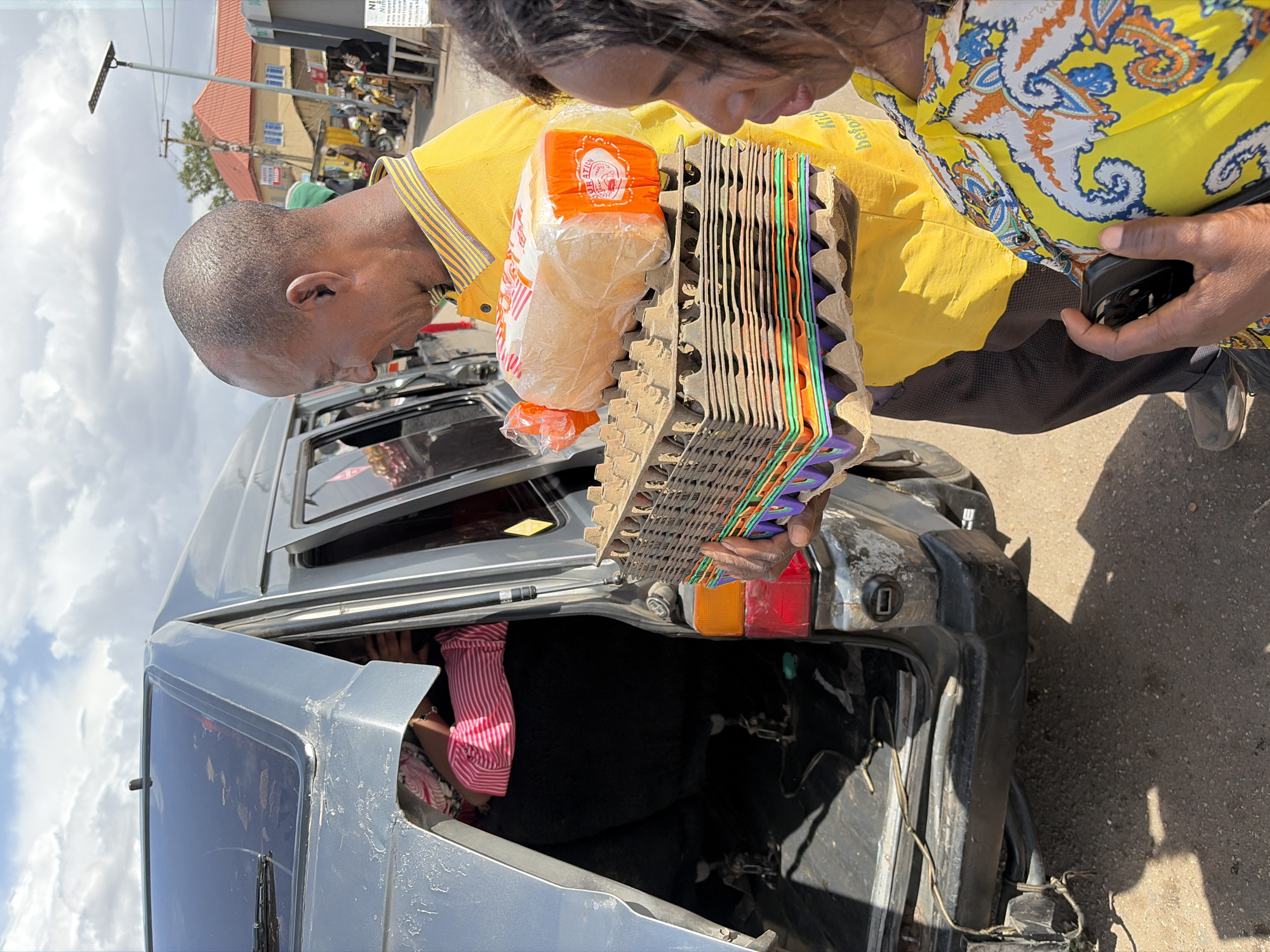 The image captures an outdoor scene in daylight, likely on a street or roadside in Jos, Nigeria. The sky is partly cloudy.
The primary subjects are two individuals loading items into the open trunk of a gray car. A man, seen in profile, has a closely-shaved head and wears a yellow collared shirt with striped trim. He holds a clear plastic bag containing a loaf of white bread, which has "BIG ARO" and other text on its orange packaging. Beside the bread, he carries a stack of cardboard egg crates, some of which contain colorful plastic inserts in shades of green, orange, and purple.
Next to the man, a second person, possibly female, is partially visible, also looking down at the items. She wears a yellow shirt with a distinctive blue, orange, and white paisley pattern and holds a black smartphone in her hand.
The gray car shows signs of wear, including dirt and scratches. Its trunk is open, and a person wearing a pink and white striped shirt is partially visible inside, suggesting they might be arranging items.
In the background, several buildings with orange roofs are visible, along with a solar-powered street light. The scene depicts a routine activity, possibly involving the transport or delivery of bread and eggs.