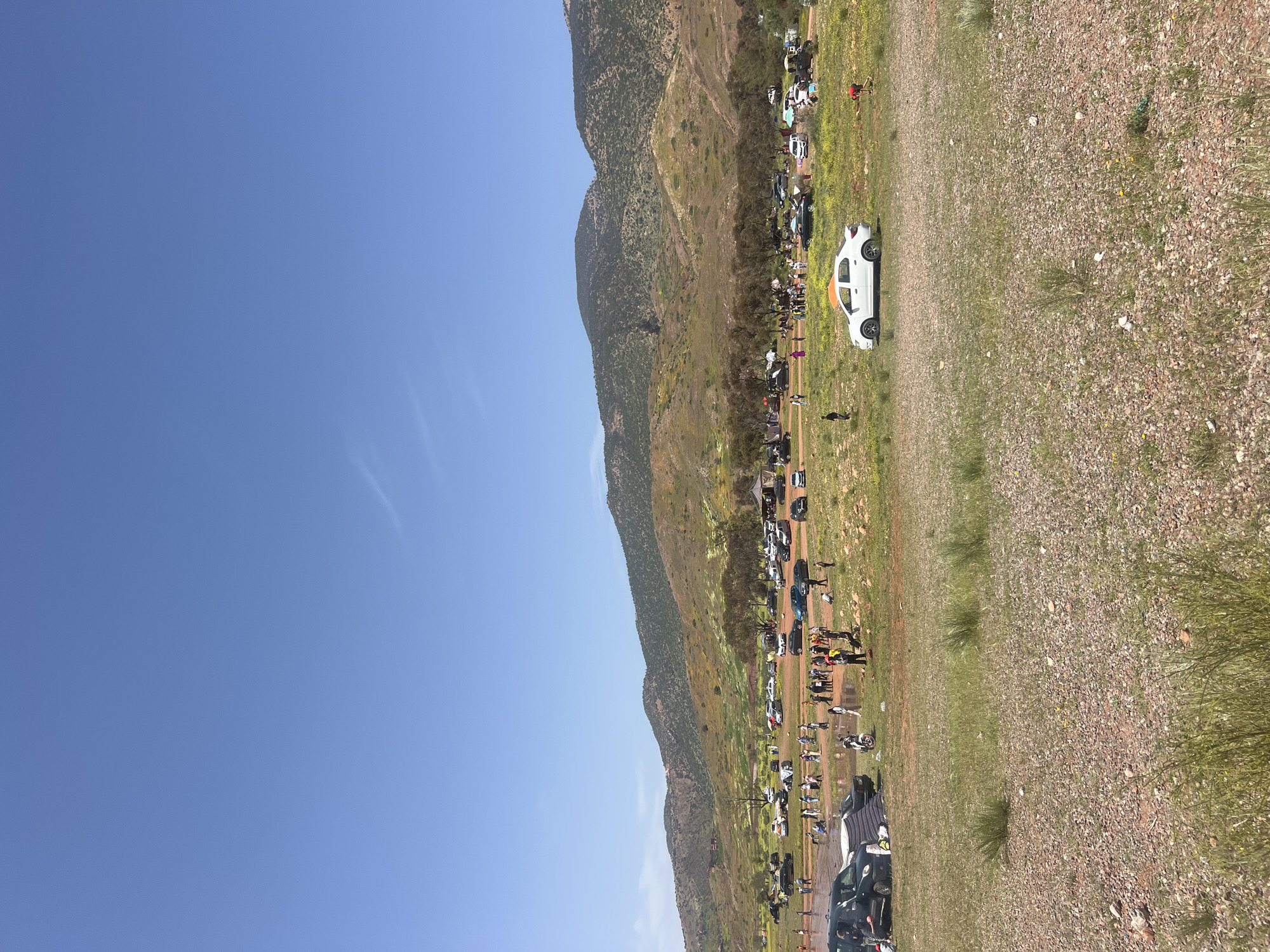 The image shows a gathering of people and vehicles in a natural environment. A mountain range rises along the left edge of the frame. A road, lined with parked cars and pedestrians, stretches between the mountains and a grassy, gravelly area. A white car with an orange roof is prominently visible in the foreground. The sky is clear and blue. This image was taken in Bin El Ouidane, Morocco.