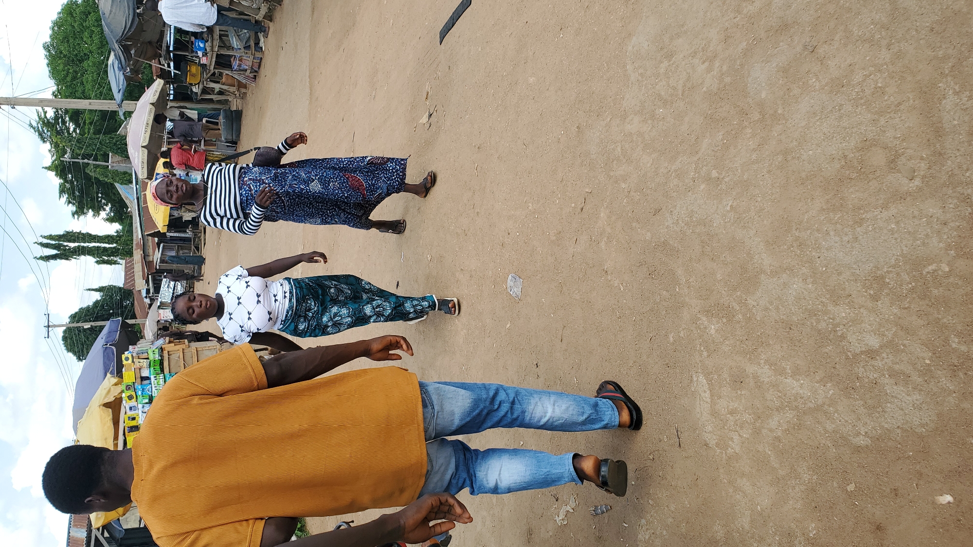 In Jalingo, Nigeria, three individuals are walking across a dirt road in a market setting. On the left, a person wearing an orange, ribbed t-shirt and light blue jeans walks away from the camera, their back to the viewer. They are wearing black sandals with red and green stripes. In the center, a young woman in a white t-shirt adorned with a bow pattern and a dark blue and green patterned skirt walks towards the camera, her eyes closed. She is wearing black sandals. To the right of her, a woman with a red headscarf, wearing a black and white striped t-shirt and a dark blue patterned skirt, walks towards the camera. She has a dark bag slung over her left shoulder and is wearing dark sandals. Behind the people, stalls with awnings are visible, displaying various goods, including what appears to be electronics. The environment suggests a bustling outdoor market.