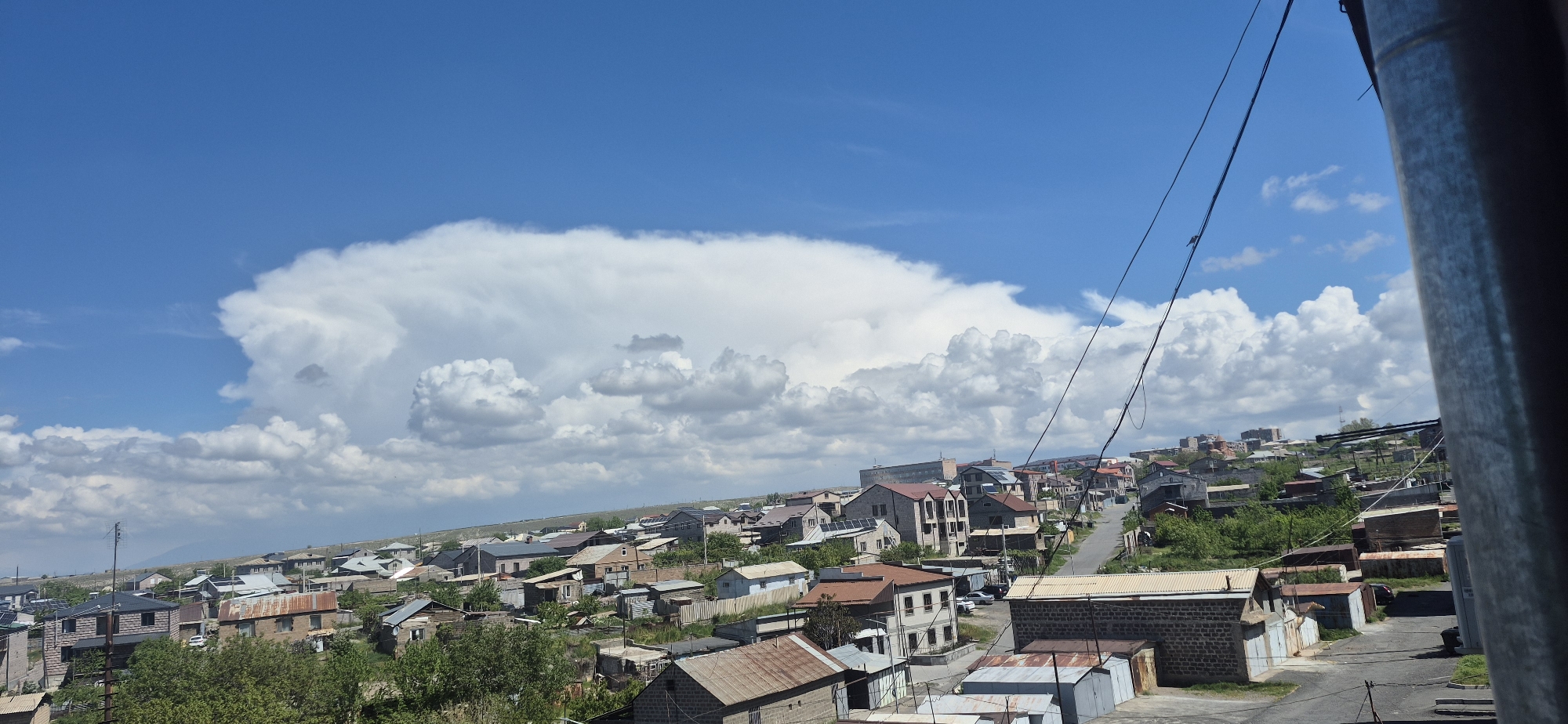 The image depicts a neighborhood in the city of Unknown, Unknown, under a bright blue sky with scattered white clouds. A large cumulus cloud formation dominates the left side of the sky. The neighborhood consists of a mix of single-story and multi-story buildings with various roof types, including metal and tile. Some buildings have visible signs of weathering and aging. Roads and paths weave through the neighborhood. Power lines run across the upper right portion of the image. A cylindrical object is partially visible on the right edge of the frame. The overall scene suggests a residential area with a mix of older and newer construction.