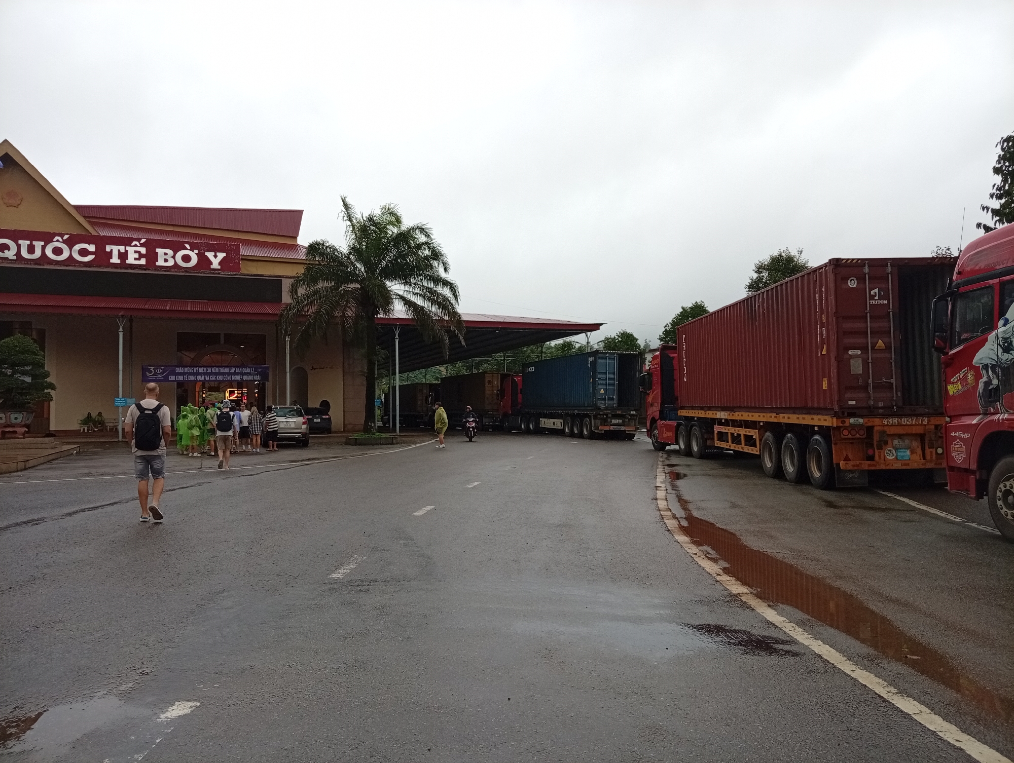 The image shows the Bo Y International Border Gate in Bo Y, Vietnam. A man with a backpack walks away from the camera on a wet asphalt road. In the background, a building with red roofing and signage that reads "QUỐC TẾ BỜ Y" is visible. Several people are gathered near the entrance of the building, some wearing bright green ponchos. A line of large trucks, including container trucks, are parked or moving along the road. The sky is overcast, and puddles of water are present on the road surface, indicating recent rain. Trees and vegetation are visible around the border gate area.