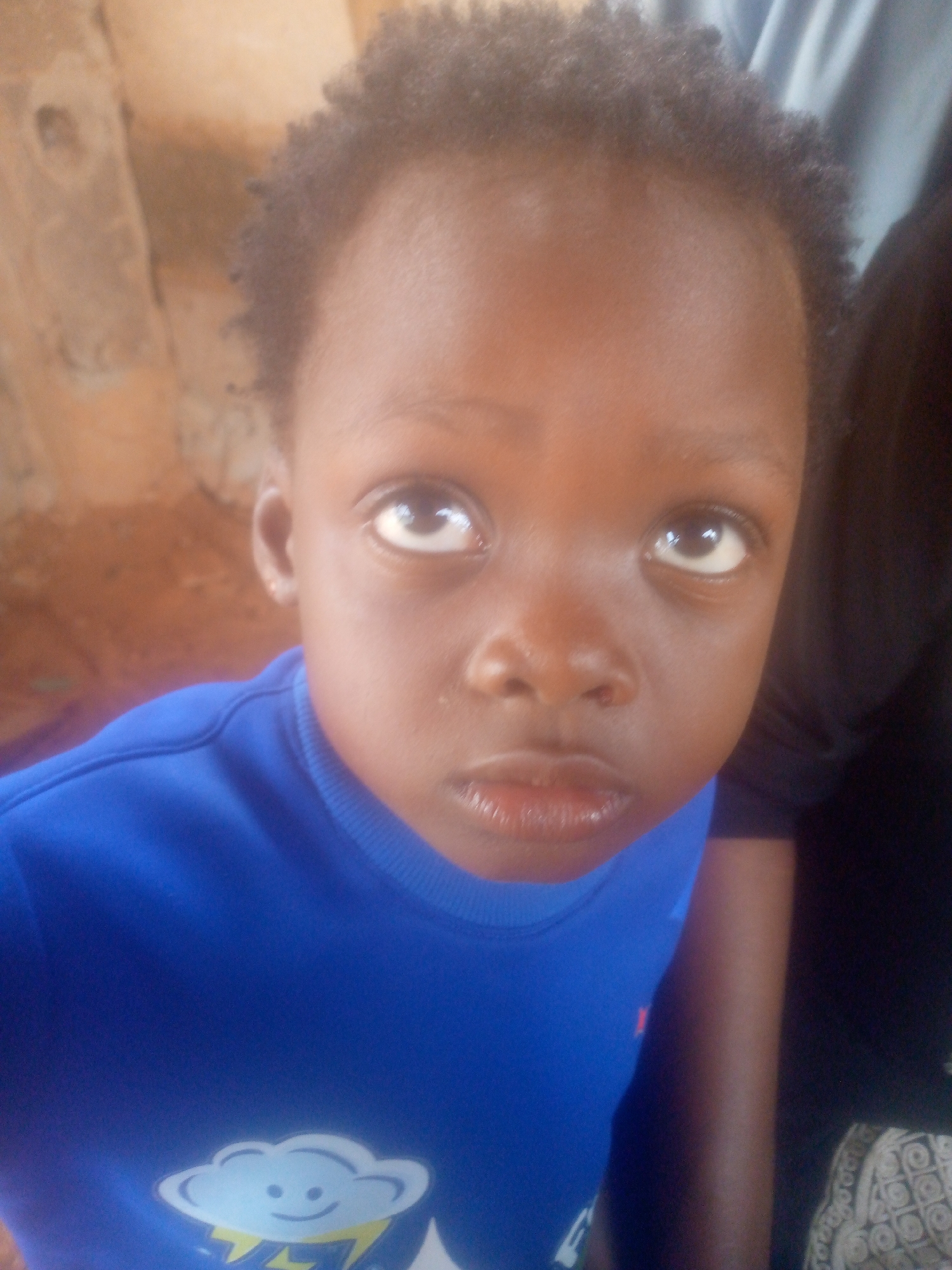 A young child with dark skin and short, dark hair looks upward with a slightly pensive expression. The child is wearing a bright blue crewneck shirt with a cartoon cloud design on the front. The background appears to be a room with blurred interior walls. Part of another person is visible to the right, wearing dark clothing. The location is specified as Ikorodu, Nigeria.