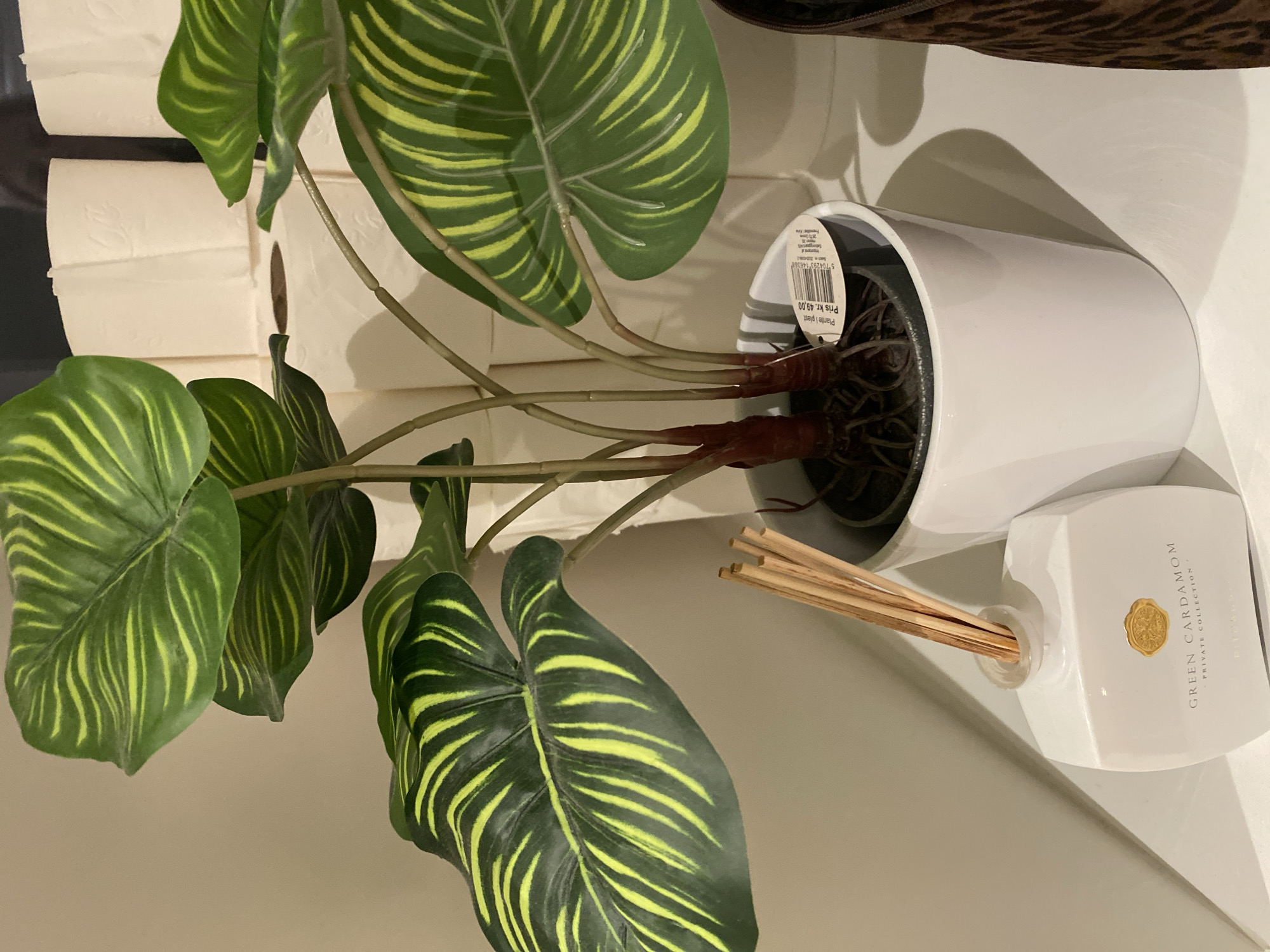 An indoor artificial plant with large green and yellow striped leaves stands in a white pot on a white shelf. To the left of the plant is a white diffuser bottle with reeds, labeled "GREEN CARDAMOM - PRIVATE COLLECTION". Behind the plant, several rolls of white toilet paper are stacked. To the right of the plant, a portion of a brown and black leopard-print item is visible. The scene appears to be in a residential setting, possibly a bathroom or living room, in Herning, Denmark.