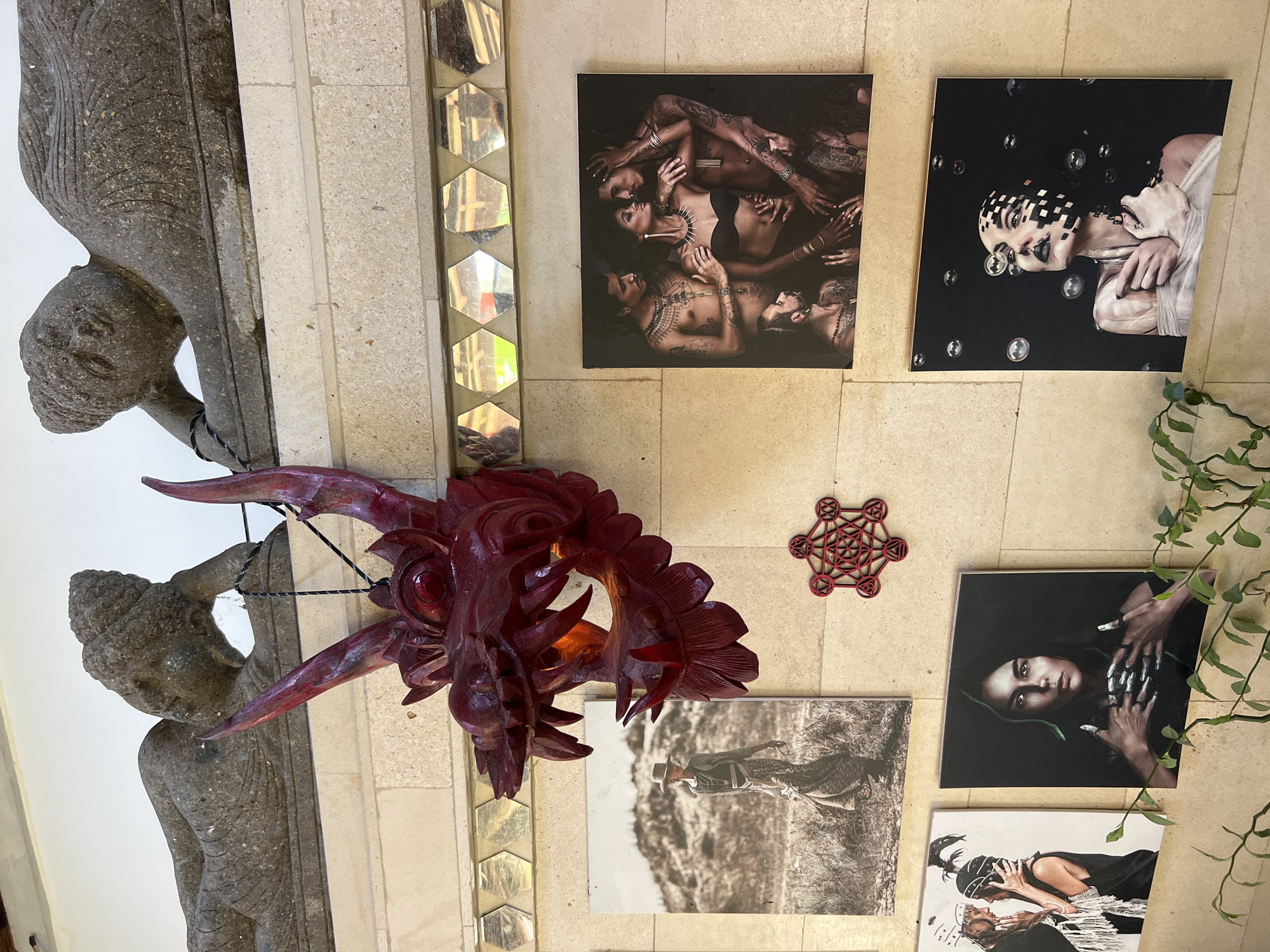 The image depicts an indoor scene in Sukawati, Indonesia. A gray stone statue of a figure supports a wall, adjacent to a strip of hexagonal mirrored tiles. A large, red, ornate dragon head with protruding horns and teeth hangs from the wall. The wall is beige and features various framed photographs: a group of tattooed people lying together, a woman with a black-and-white patterned face surrounded by bubbles, a person in a cowboy hat in a landscape, a woman with long fingernails and dark hair, and two figures embracing. Above the cowboy picture, there is a red geometric mandala-like decoration. Green leafy plants are on the right side of the photo.