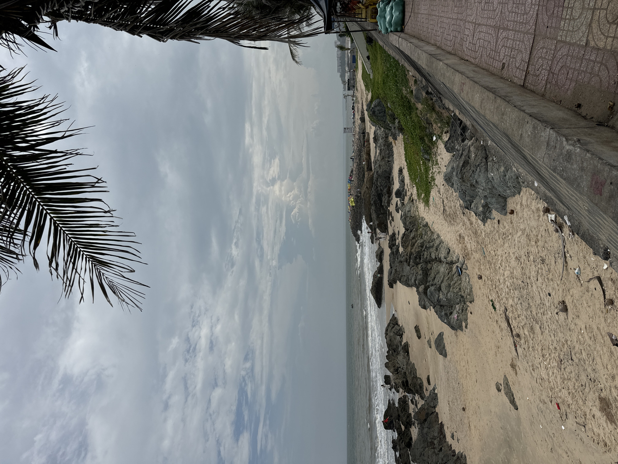 The image depicts a beachfront scene in Mũi Né, Vietnam. The foreground features a sandy beach littered with rocks, debris, and scattered coconuts. To the right, a concrete retaining wall separates the beach from a tiled walkway, upon which are several large, green cylindrical objects. Beyond the retaining wall, a dark structure with a canvas roof is visible. The middle ground displays the ocean with moderate waves crashing onto a rocky shoreline. Several people are present on the beach and in the water, some sitting on rocks, others appearing to walk along the shore. In the distance, the coastline stretches out, with scattered structures and vegetation. The sky is overcast with scattered clouds. The overall scene conveys a somewhat unkempt, natural beach environment.