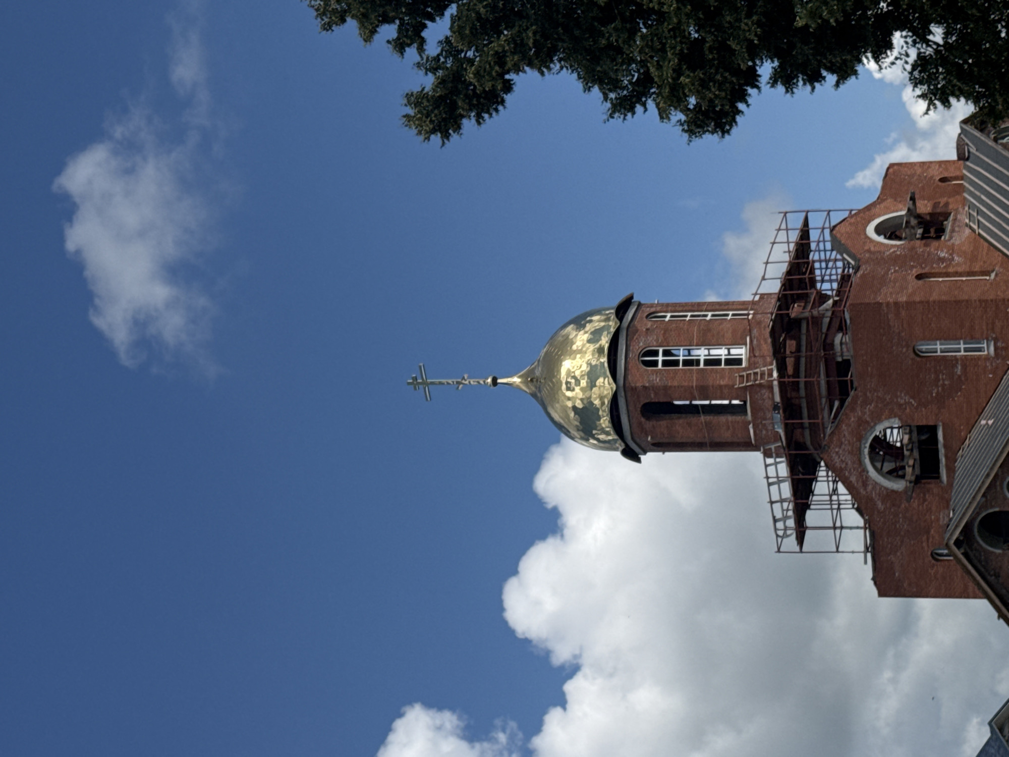 A red brick church with a golden dome topped with a cross is under construction against a blue sky with white clouds. Scaffolding surrounds the upper portion of the church tower. A large, dark green tree is on the right side of the image, partially obscuring the sky and the church. The scene appears to be outdoors in a relatively open area. There are no people visible in the image.