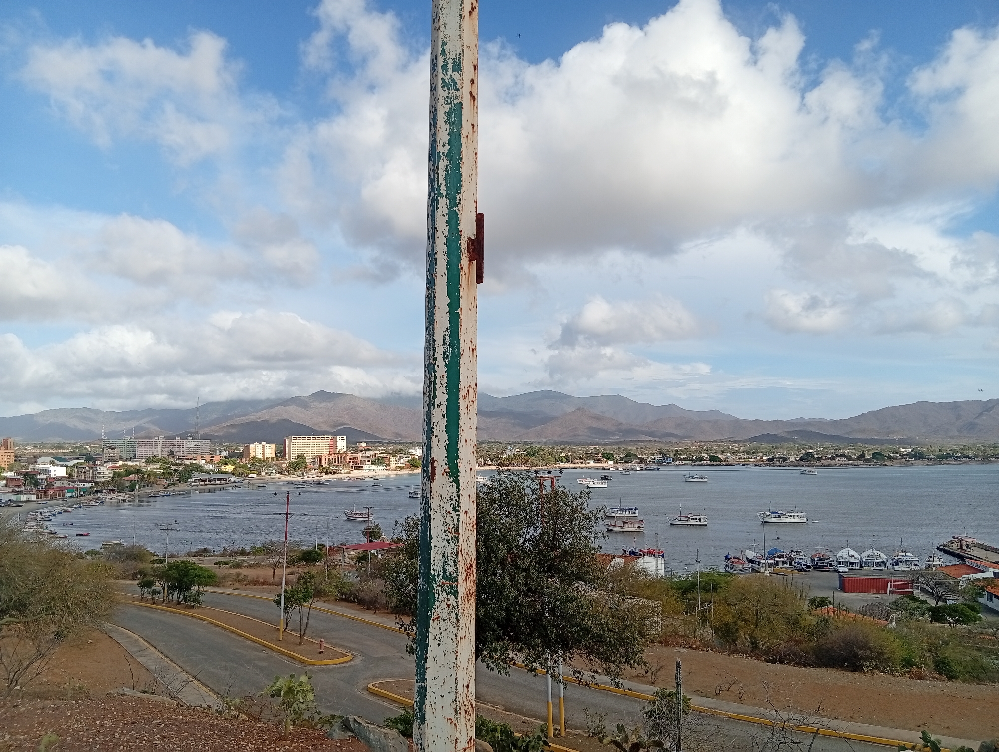The image presents a panoramic view of Juan Griego, Venezuela, from an elevated vantage point. Dominating the foreground is a weathered, cylindrical pole, its paint peeling, obstructing the view. Below the viewpoint, a winding road descends, lined with sparsely distributed vegetation and utility poles. Beyond the road, a body of water stretches out, dotted with numerous boats and extending towards the coastline. The cityscape of Juan Griego is visible along the shore, with buildings of varying heights interspersed with trees. The backdrop is formed by a series of mountains, their peaks silhouetted against a sky filled with a mix of blue and billowing white clouds.