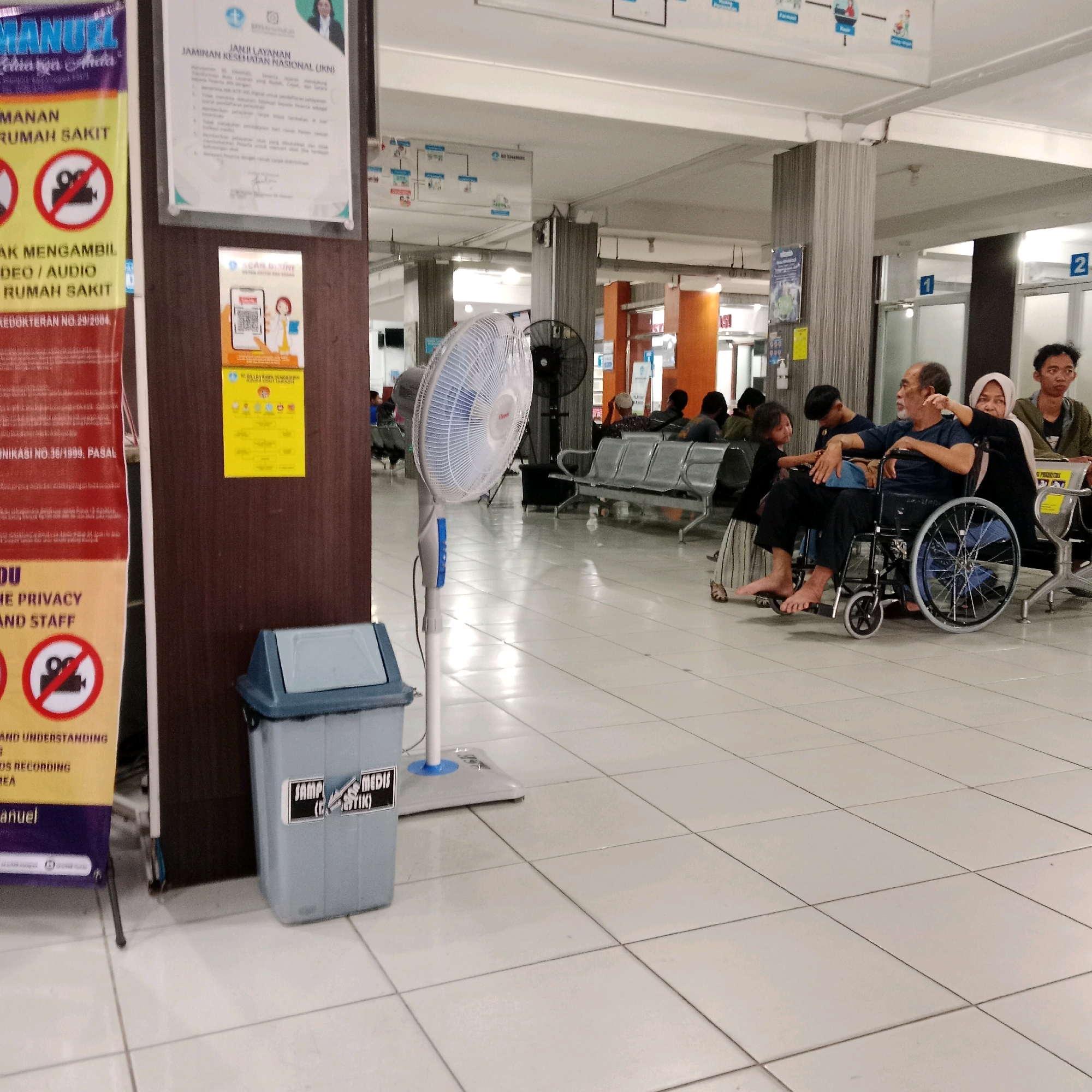 The image depicts a waiting area within a building, possibly a hospital or clinic, in Panggisari, Indonesia. In the foreground, a white floor fan stands prominently, angled to the left. To the left of the fan, a gray trash bin with a blue lid is visible, bearing a black and white label that reads "SAMPAH MEDIS (BIOTIK)".
A tall, dark brown wooden pillar is positioned between the trash bin and a red banner on the far left. This banner features text in yellow and white, including a prohibition symbol with a camera icon, indicating no recording is allowed.
In the background, a group of people are seated on metal benches. One individual is in a wheelchair, accompanied by a woman in a headscarf. Several other individuals are also present, some standing and some seated, their faces and attire suggesting a casual public gathering. Various signs and posters are mounted on the walls and pillars, some displaying text and graphics. Two numbered doors, labeled "1" and "2", are visible in the distance. The overall environment appears to be indoors, with tiled flooring and overhead lighting.