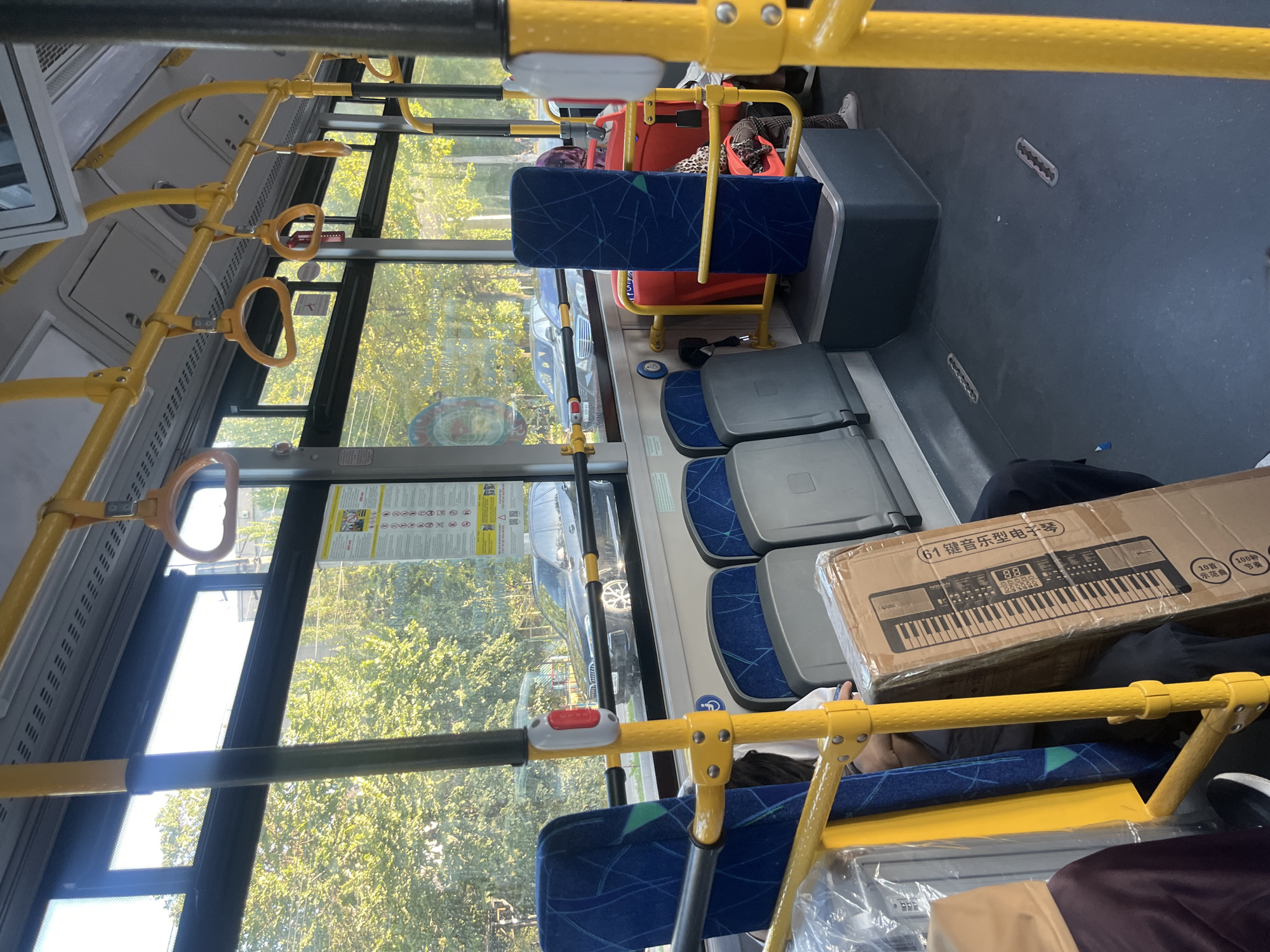 The image shows the interior of a public bus in Bishkek, Kyrgyzstan. The bus is equipped with yellow handrails and blue seats. Outside the window, trees and cars are visible, indicating an urban environment. A large cardboard box containing a keyboard is placed on the floor near the seats. Several passengers are present, though their faces are not clearly visible. The bus appears to be in motion, as suggested by the slightly blurred background. The lighting suggests it is daytime, likely with sunlight filtering through the windows. The scene depicts everyday public transportation in Bishkek.