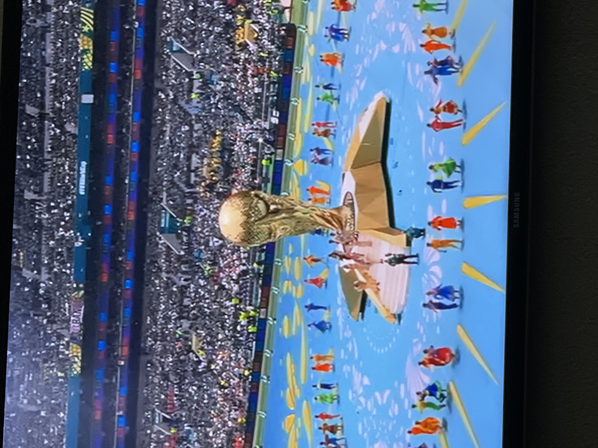 The image shows a large stadium filled with spectators. In the center of the field, a stage is set up with the FIFA World Cup trophy prominently displayed. Several individuals are on the stage and on the field in front of it, some appearing to be performing or dancing. The field itself is decorated with a blue surface and patterned designs. In the background, the stadium seating is packed with people. The overall scene suggests a sporting event ceremony or celebration. The location of the city of Moscow, Russia, is not depicted or implied in this image.