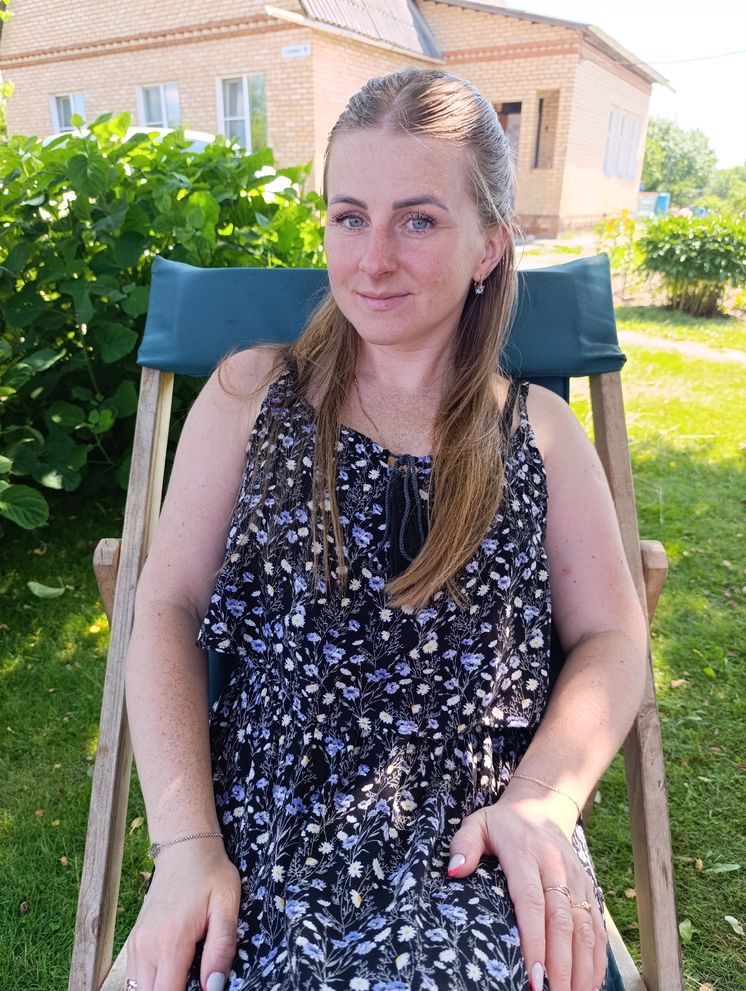 A medium shot shows a woman sitting in a wooden lounge chair outdoors. She has blonde hair pulled back in a ponytail and is wearing a black floral dress. She has blue eyes and freckles on her face and arms. The background shows a brick building and lush green foliage. The lighting suggests it is daytime and sunny.