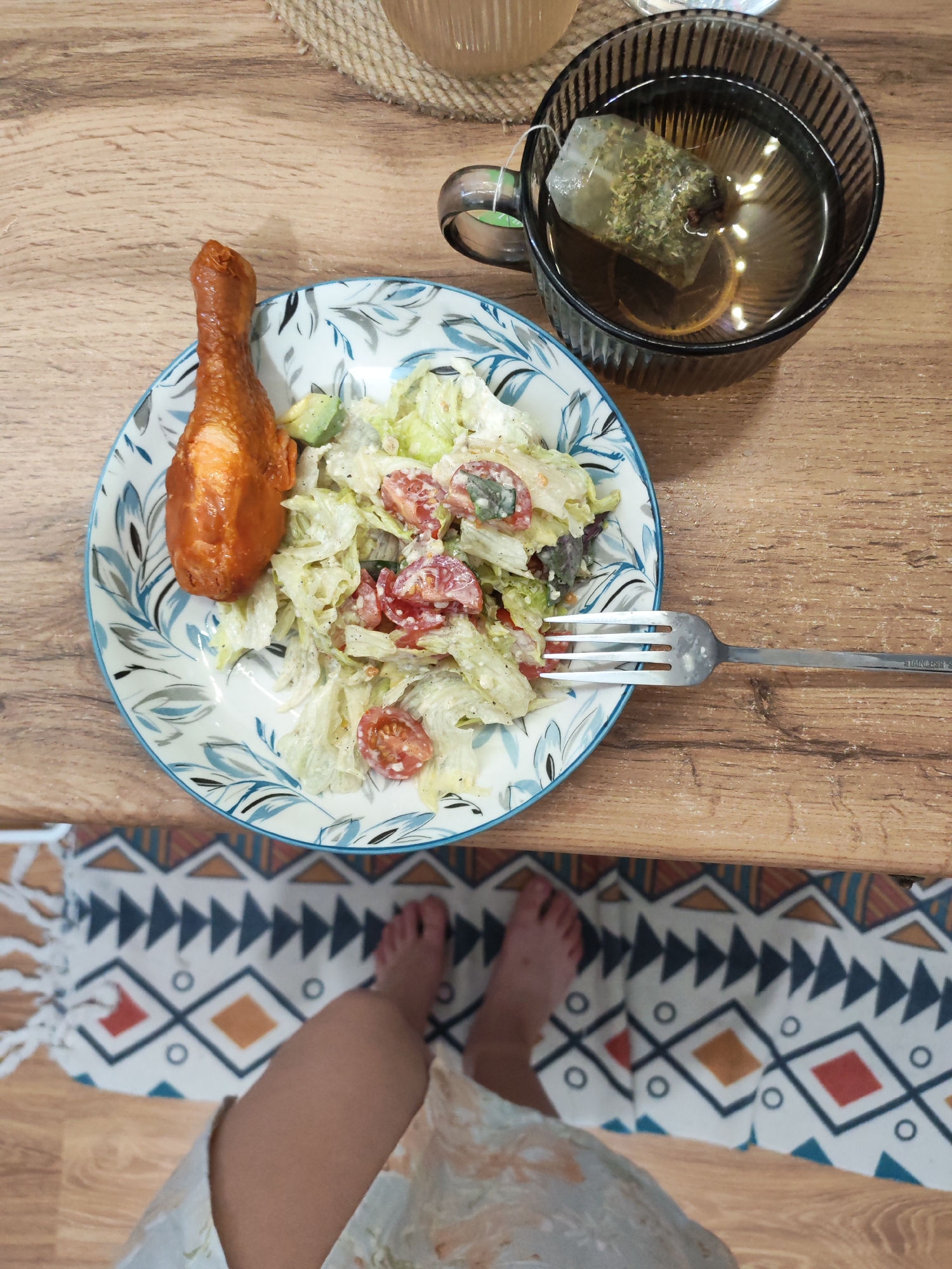 A top-down shot shows a meal laid out on a wooden table, suggesting a home dining setting. A plate with a chicken drumstick and a salad, accompanied by a mug of tea and a fork, are arranged on the table. The meal appears to be a light lunch or dinner, with the salad consisting of lettuce, cherry tomatoes, and a creamy dressing. The chicken drumstick is golden brown and looks freshly cooked. The mug of tea has a tea bag steeped in it, indicating a warm beverage to accompany the meal. The background shows a glimpse of a patterned rug and a person's legs, suggesting the meal is being enjoyed by someone sitting at the table. The overall impression is one of a simple, home-cooked meal. The image does not contain any information that would allow for its location to be specified to Izhevsk, Russia.