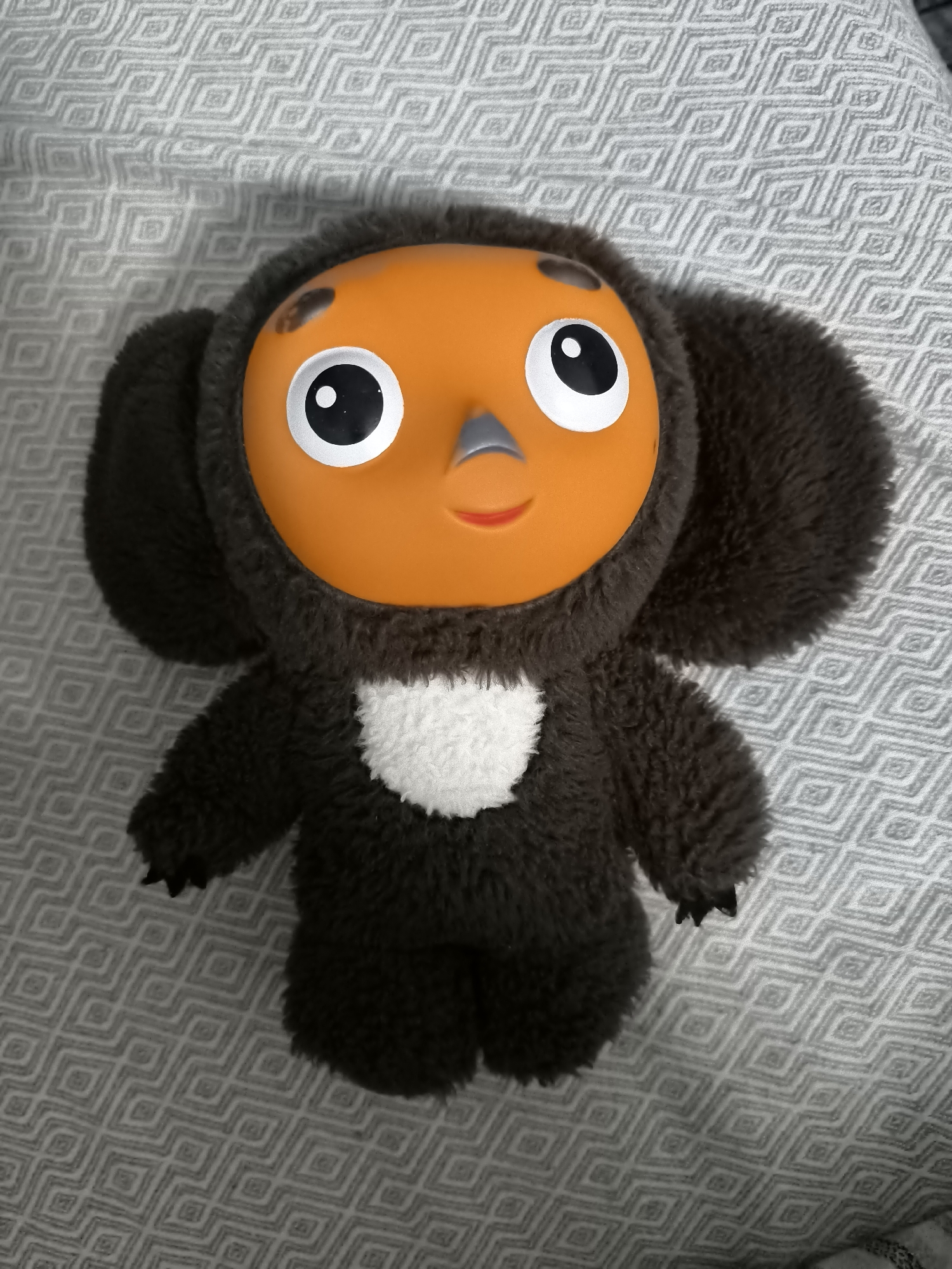 A plush toy, resembling the character Cheburashka, is positioned centrally on a textured gray surface, likely a blanket or bedspread with a diamond-shaped pattern. The toy is dark brown with large, round, furry ears and a distinctive orange face. It has prominent black and white circular eyes, a small gray nose, and a curved orange mouth. A white fluffy patch is visible on its chest. The toy is upright and appears to be placed, not in motion. There are no people or other objects visible in the image. The setting does not provide any specific indicators to place it within the city of Kokand, Uzbekistan.