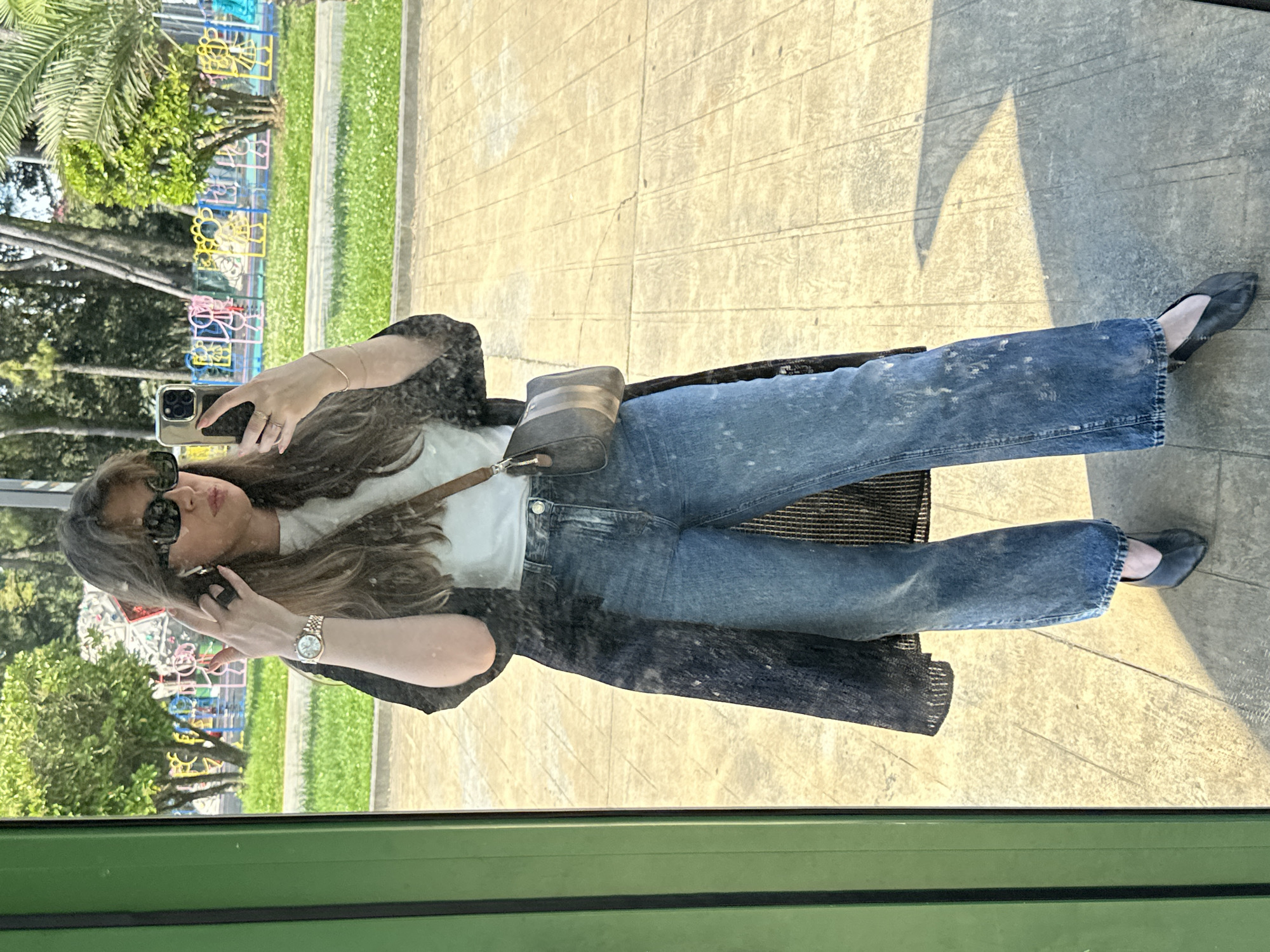 A woman takes a mirror selfie in Batumi, Georgia. She wears jeans, a white t-shirt, a dark cardigan, sunglasses, and flat shoes. She has a small bag over her shoulder and is holding her phone. In the background, there's a park with green trees and colorful metal structures characteristic of Batumi's urban landscaping. The light suggests it's a sunny day. The frame of the mirror is green, indicating an enclosed structure, possibly a public bus stop or a similar shelter. The ground is paved.