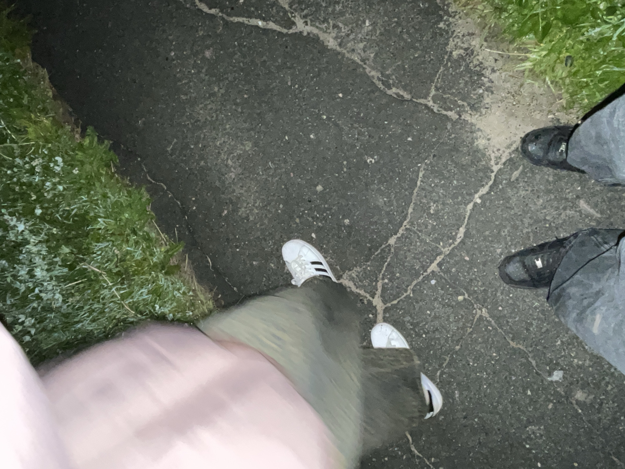 The image provides an overhead view of the lower bodies of two individuals on a cracked asphalt path next to green grass. The setting appears to be at twilight or night due to the low light conditions.
On the left, one person is visible, wearing a light-colored (possibly pink) top or jacket over a green skirt or trousers, and white Adidas sneakers with black stripes. This person's lower half is blurred, suggesting movement, possibly walking or the image was taken while in motion.
On the right, the second person wears grey pants and black sneakers. A small, light-colored logo, possibly resembling a "J", is visible on the side of one of the black sneakers.
The ground consists of dark, speckled asphalt, extensively cracked with lighter lines. Along the edges of the asphalt, vibrant green grass is visible, appearing somewhat damp or dewy. The scene is an outdoor setting, such as a path or sidewalk, located in Красные Баки, Russia.