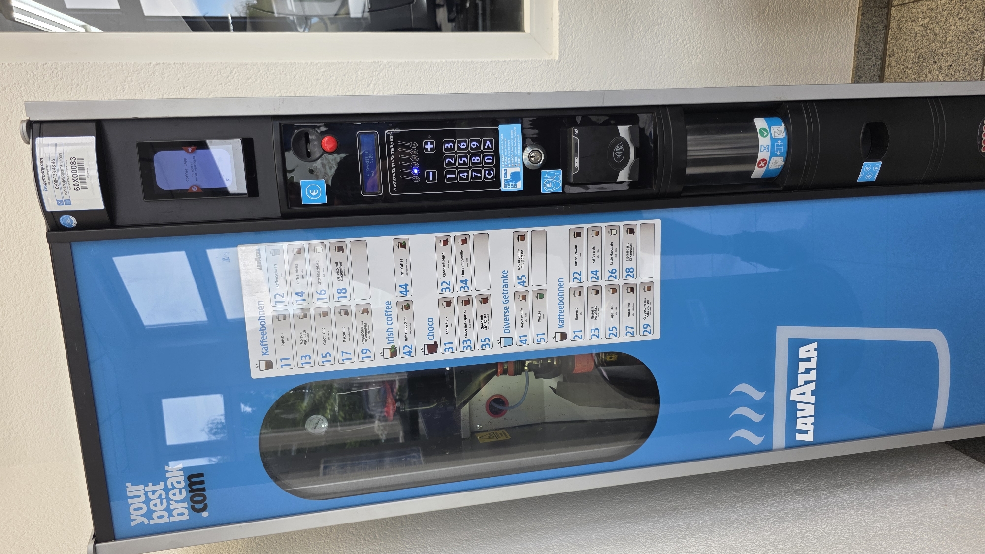 An exterior, eye-level, full shot captures a bright blue Lavazza vending machine situated in front of a pale beige wall. The vending machine is predominantly blue with white lettering that reads "your best break .com" on the upper left side. The machine features a glass door on the left, revealing internal components including a metal frame and a small gauge. The right side of the machine is black and houses a touchscreen display, a payment terminal for coins and cards, and a keypad with numerical buttons and symbols. A selection of beverage options, including various coffee drinks and hot chocolate, are listed on the front of the machine with corresponding numbers. The background is blurred, but suggests an indoor environment with windows and a glimpse of a white wall. The lighting is bright and even, highlighting the details of the vending machine. There are no people or animals present in the image.