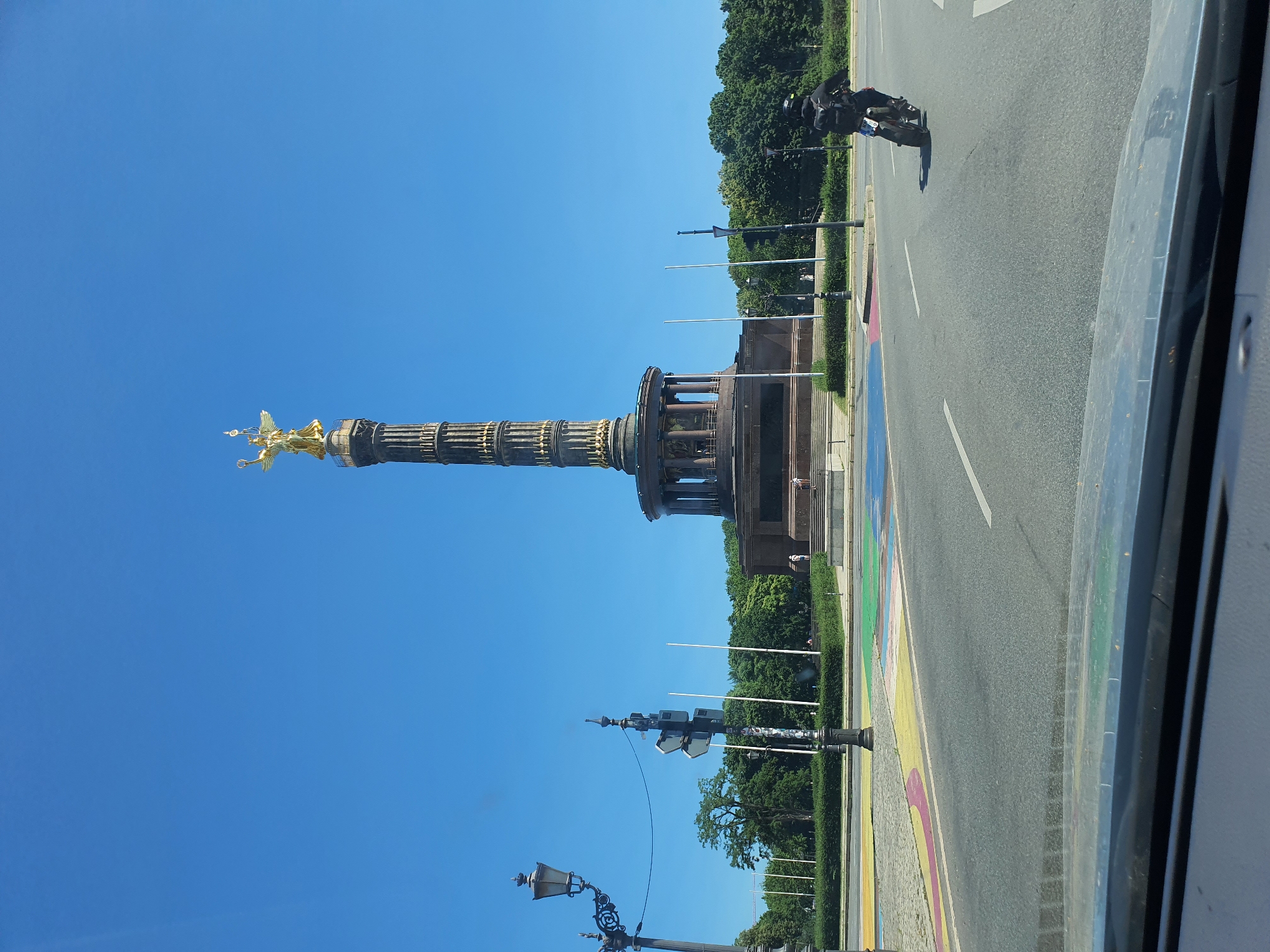 A wide-angle shot captures the Victory Column in Berlin, Germany, on a clear, sunny day. The iconic monument, topped with a golden statue of Victoria, stands tall against a bright blue sky. The column's detailed structure, with its ornate carvings and tiered design, is clearly visible.
In the foreground, a person rides a motorcycle on a street painted with colorful stripes, resembling a rainbow. The motorcycle has a rider wearing a helmet and dark clothing. The street is paved with asphalt, and white lines mark the traffic lanes.
Beyond the monument, lush green trees form a dense line, suggesting a park or well-maintained urban landscape. Lampposts and traffic signals are also visible along the street. The scene appears to be during daylight hours, with the sun casting distinct shadows. The overall impression is one of a vibrant and active urban environment in Berlin.