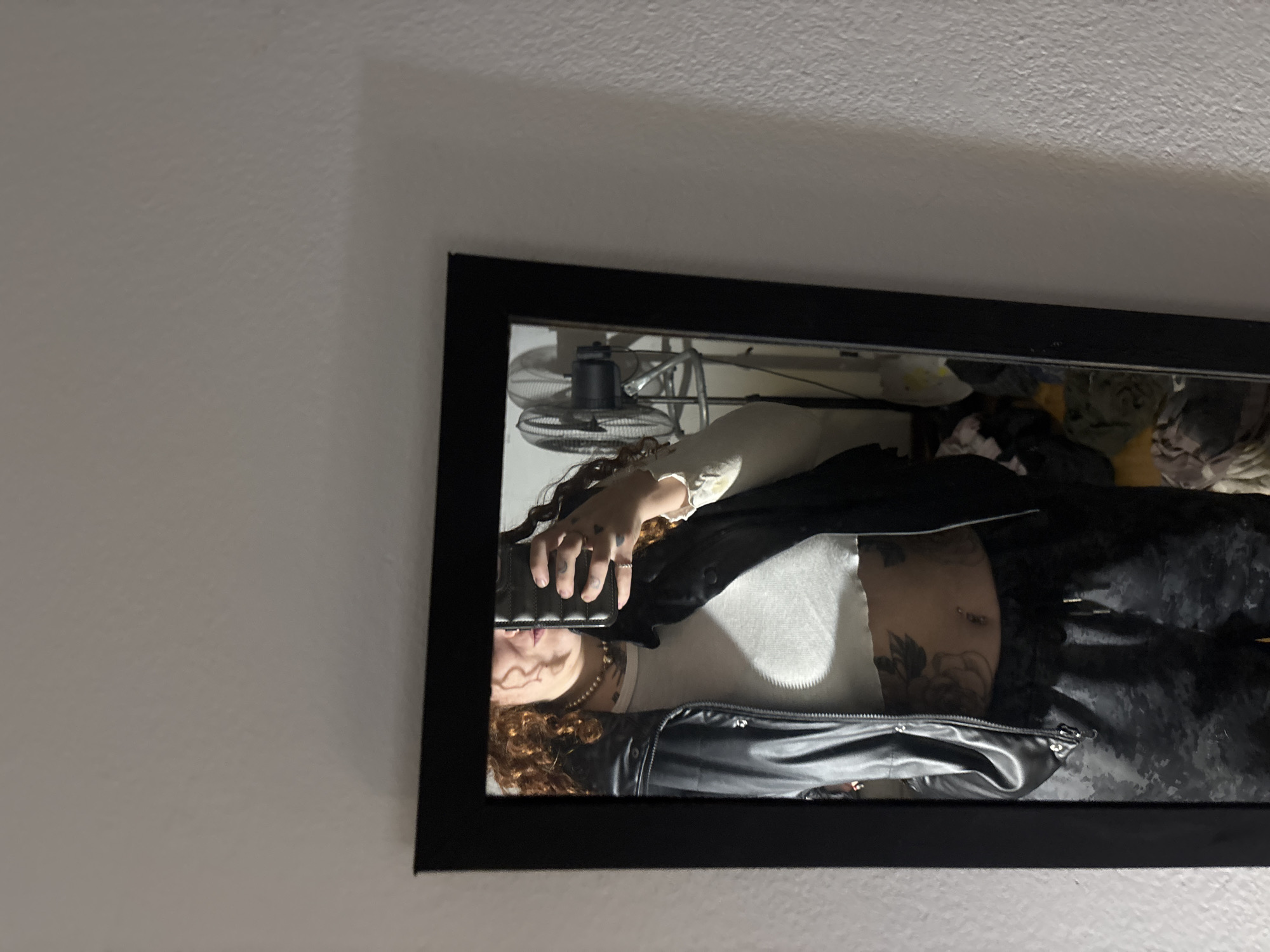 A mirror, framed in black, is positioned vertically against a plain, light-colored wall. Reflected within the mirror is a person, likely standing indoors. The person has curly, reddish-brown hair and is holding a black rectangular object, presumably a phone, in their hand, partially obscuring their face.
They are wearing a white long-sleeved top that exposes their midriff, over which a black jacket, possibly made of leather or a similar material, is layered. The lower body is dressed in dark, patterned pants, which appear to have a camouflage-like design. Notable details on the person include a belly button piercing and several tattoos, including floral designs on their stomach and smaller symbols on their fingers. Multiple rings are worn on their fingers, and a beaded necklace adorns their neck.
In the background of the reflection, behind the person, a metal floor or desk fan is visible. The lighting suggests an indoor environment, possibly illuminated by artificial light, creating some shadows. The scene appears casual, capturing a moment of the person looking at their reflection or taking a mirror selfie. No visible text is clearly discernible within the image. The image was taken in Buenos Aires, Argentina.