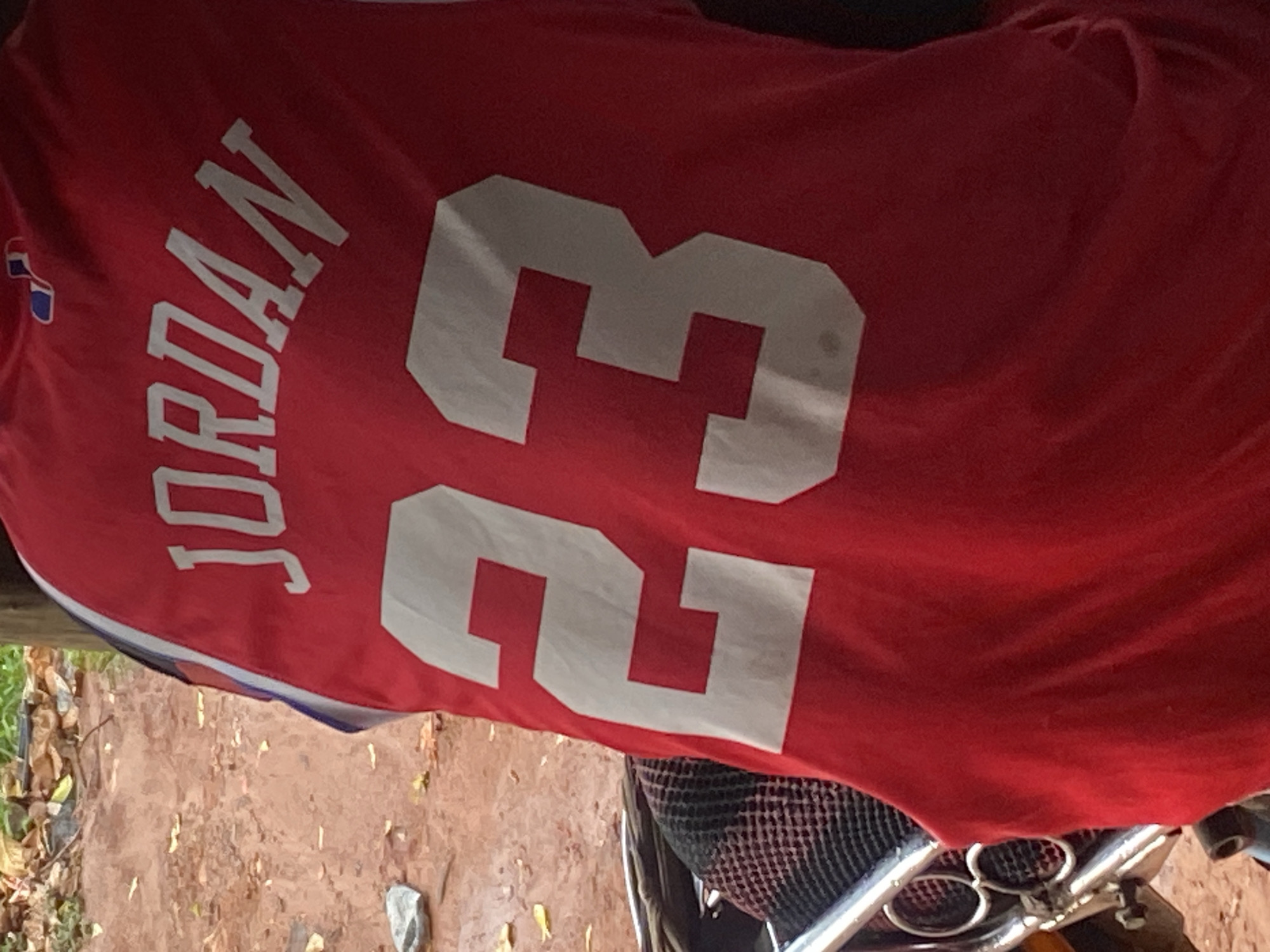 A red sports jersey, emblazoned with the name "JORDAN" printed vertically in white capital letters and the large white number "23" horizontally, dominates the frame. The jersey features a subtle blue and white trim along its visible edge, and a small, partially obscured blue and red emblem or logo can be seen on the upper left side, near the 'J' in Jordan.
The jersey appears to be draped over or resting on an object, possibly a vehicle such as a motorcycle or scooter, indicated by metallic components and black mesh material visible in the bottom right corner of the image. Below the jersey and vehicle, the ground is an earthy brown, suggesting an unpaved surface, scattered with dried leaves and small rocks.
The setting is outdoors, likely a casual street or yard area, characteristic of an everyday environment in Auchi, Nigeria. The lighting indicates it is daytime. No individuals are visible, and no specific activity is occurring. The image is presented with a rotated perspective.