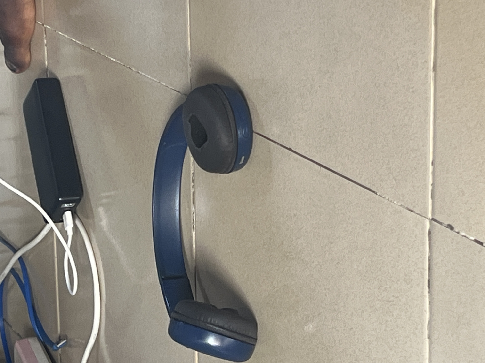 The image shows a close-up of blue headphones hanging on a tiled wall in Jauro Yinu, Nigeria. The headphones are positioned so that one earcup and part of the headband are visible. The wall is tiled with light-colored, textured tiles, and there are visible grout lines.
To the left of the headphones, a black power bank is plugged into a white cable. Other wires, including a blue one, are also visible near the power bank, suggesting a charging or connectivity setup. A portion of a finger or thumb is visible in the top left corner, indicating a person is present, possibly holding the headphones or interacting with the devices.
The lighting suggests it is daytime. The overall scene appears to be indoors, given the tiled wall and the presence of electronic devices. There are no strong indicators of specific weather conditions or time of day beyond daylight. The focus is on the headphones and the adjacent electronic accessories. No text is visible in the image.