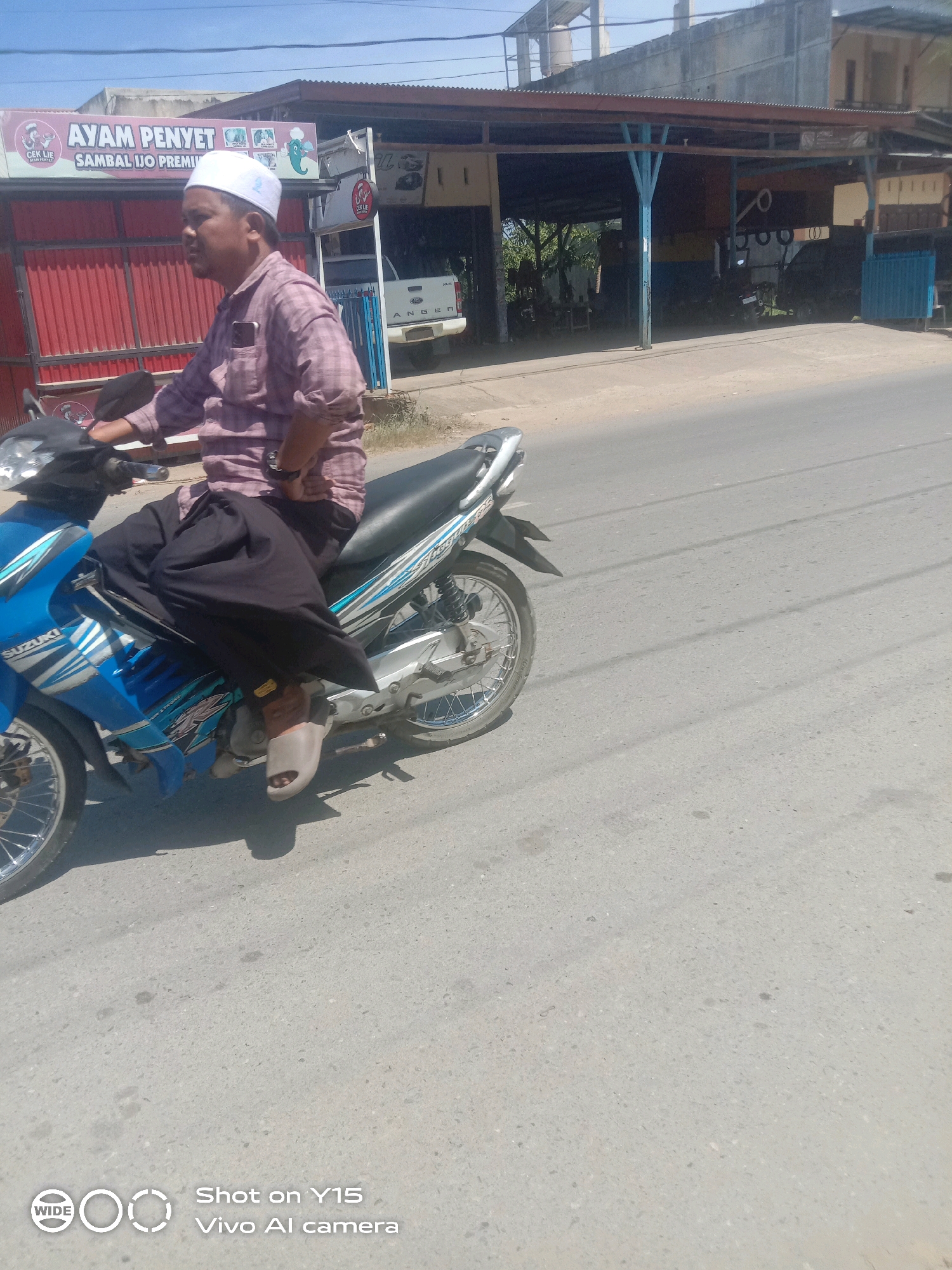 A man wearing a white peci and a maroon and white checkered long-sleeved shirt is riding a blue and silver Suzuki motorcycle on a paved road. He is also wearing black pants and light-colored sandals. The motorcycle has a black seat and a silver exhaust. In the background, there is a building with a red corrugated metal facade and a brown roof supported by blue posts. A sign above the building reads "AYAM PENYET SAMBAL IJO PREMI". A white pickup truck is parked behind the building. The sky is clear and blue, and the scene appears to be in an urban or semi-urban area. The image also contains a watermark in the lower left corner that reads "WIDE Shot on Y15 Vivo AI camera".