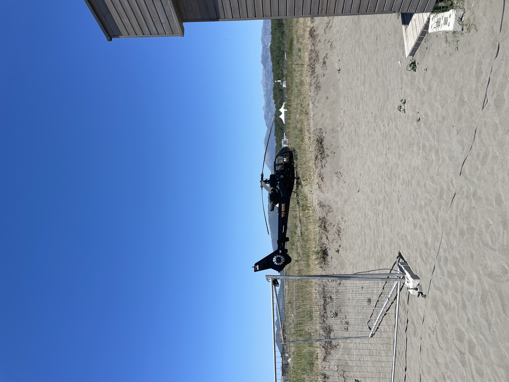 A black helicopter with the registration "YU-MNE" is parked on a sandy beach in Donji Štoj, Montenegro. The helicopter is positioned in the mid-ground of the image, with its tail rotor and tail boom visible. In front of the helicopter, there is a temporary metal fence. The beach is covered in sand and sparse vegetation. In the background, there are mountains covered with trees, and a white tent can be seen in the distance. To the right of the image, part of a wooden structure, possibly a beach hut or cafe, is visible. The sky is clear and blue.