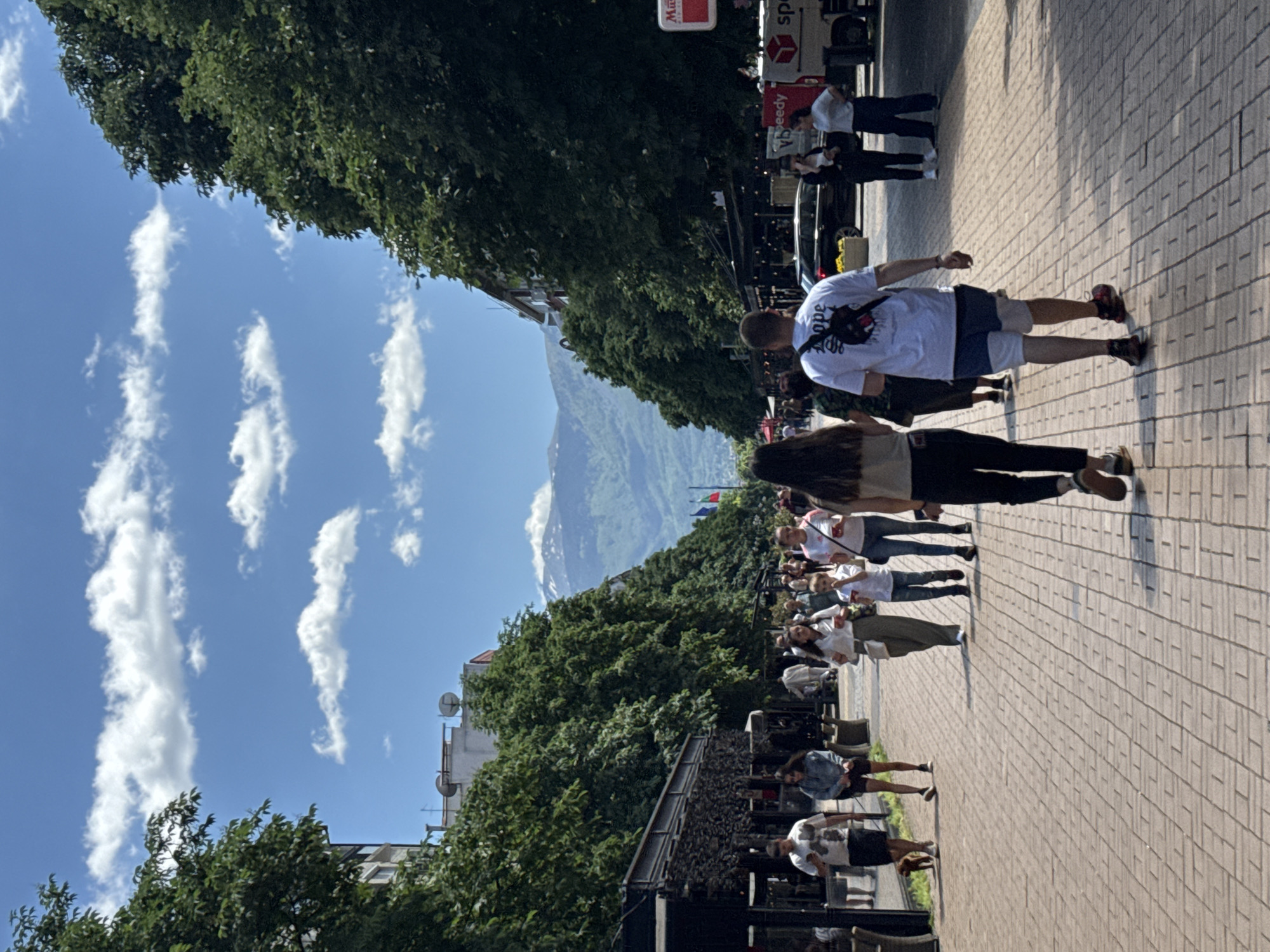 The image depicts a street scene in Sofia, Bulgaria, characterized by a sunny, clear sky with elongated, wispy clouds. The scene is populated by numerous individuals walking along a brick-paved street. On the right, a man with short hair and a white shirt is walking away from the camera. A woman with long dark hair walks beside him. Further along the street, a group of people are walking in the same direction. On the left side of the street, tables and chairs suggest outdoor dining areas adjacent to commercial buildings. Trees line both sides of the street, and in the background, a mountain is visible. A sign of a truck carrying a package can be seen in the background.