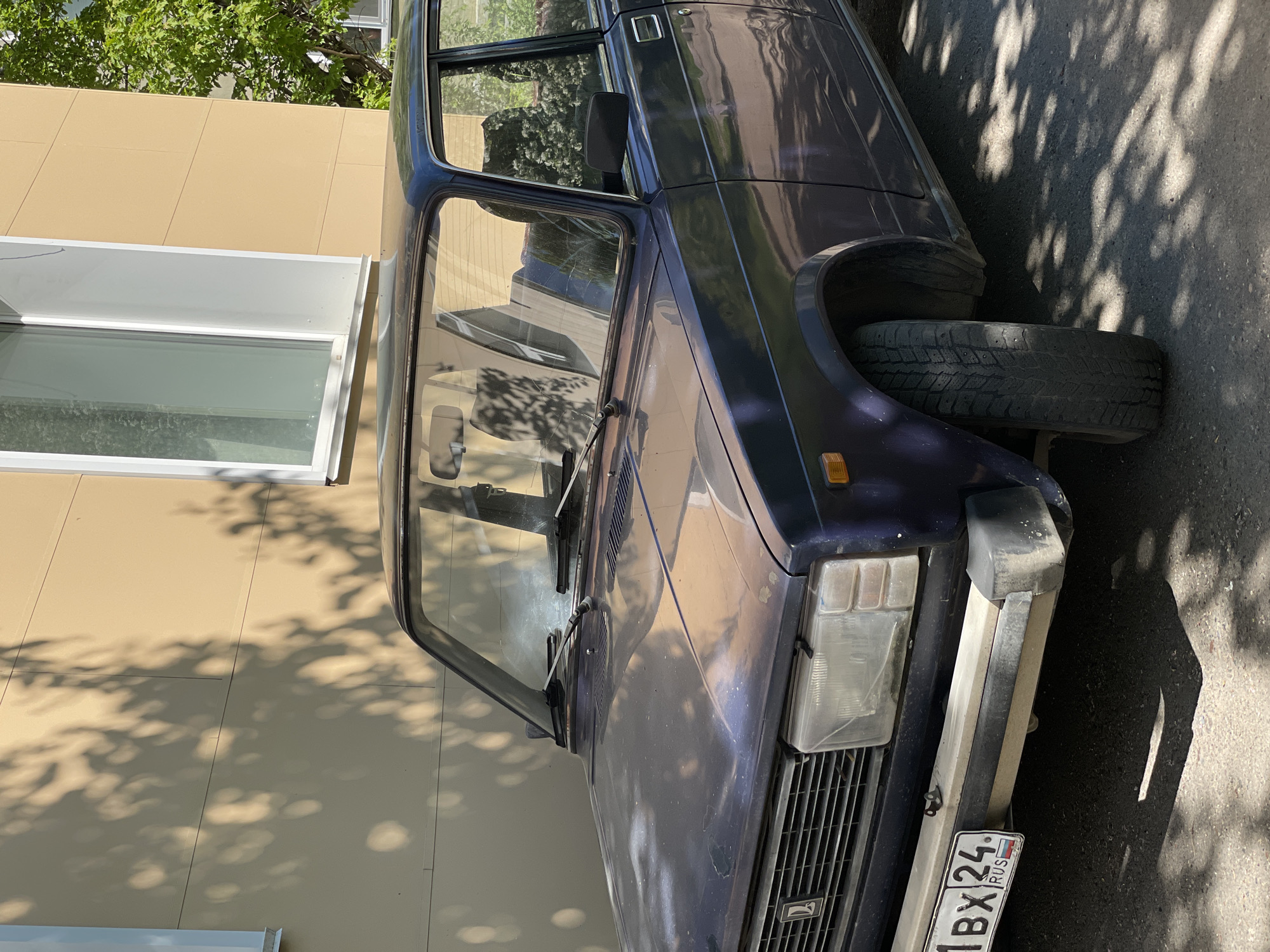 A dark blue, older model sedan car is parked on an asphalt surface next to a light brown building under bright, direct sunlight in Zheleznogorsk, Russia. The car's front is visible, featuring a rectangular headlight on the right, a gridded front grille, and a light-colored bumper with a darker protective strip. The paint appears somewhat faded and slightly dirty, indicating age and use.
A notable detail is the car's front left wheel well, which contains a loose tire leaning against the fender, suggesting the car is either undergoing maintenance or is in a state of disrepair, as a proper wheel assembly is not visible. Distinctive, dappled shadows from overhead tree foliage fall across the car's body, the building wall, and the asphalt, indicating a clear, sunny day, likely midday or afternoon.
The adjacent building has a light brown or beige facade with a large, modern white-framed window, reflecting the sky and green trees visible at the top of the frame. The scene is static, with no activity or people present beyond the parked vehicle.
Visible text on the car's front license plate clearly reads "1BX 24 RUS." The "RUS" confirms the country as Russia, and "24" is the regional code for Krasnoyarsk Krai, the administrative division where Zheleznogorsk is located.