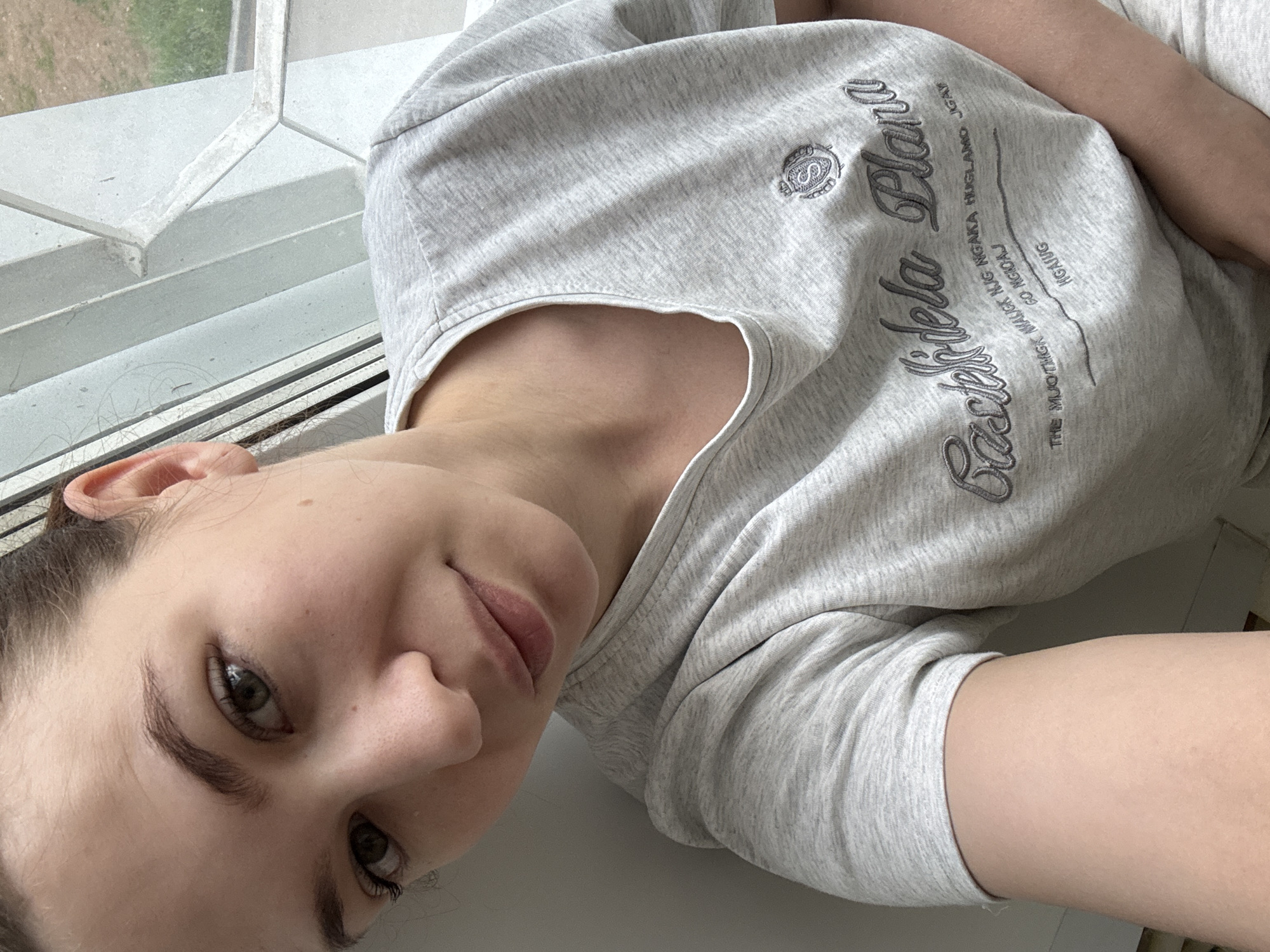 The image presents a close-up, eye-level selfie of a young adult female positioned indoors, adjacent to a window. She has fair skin, dark brown hair that appears pulled back, and green or hazel eyes with prominent, dark eyelashes and well-defined eyebrows. A small mole or blemish is visible on her right cheek. Her lips are closed, showing a slight, neutral expression.
She is wearing a light gray, heathered short-sleeved t-shirt with a rounded neckline. The t-shirt features dark gray embroidered text and a crest logo on the upper right chest. The logo is a circular emblem resembling an intertwined 'S' or '8' topped by a crown. Below this, the main text, in a script font, reads "Casteldela Plana". Further below, in a smaller sans-serif font, are several lines of text: "THE MUOTMGK MAUGC KUG NGAKA HUGLAMO JGAY", "GO NGIOAJ", and "HGAJUG".
The background to her right reveals a white, multi-paned window frame with visible dust or dirt. Through the window, green foliage and patches of brown earth are discernible, indicating an outdoor environment. A plain, light-colored wall is visible to her left. The scene is illuminated by natural light originating from the window. The scene is situated in Chaikovsky, Russia.
