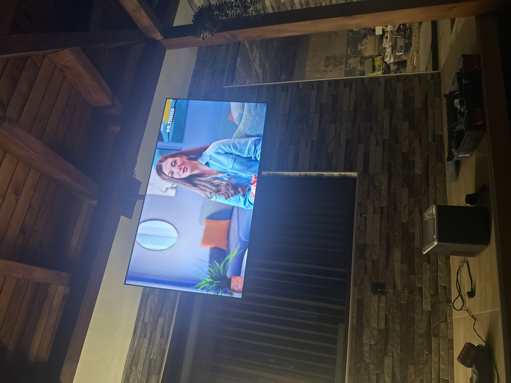 A large television screen displays a woman wearing a blue shirt, identified by the text "DR. THEISS" on the screen. The screen is mounted outdoors, projecting onto a stone wall. Below the screen, on a wooden surface, sits a black projector. A black power cord snakes across the surface towards an electrical outlet in the wall. Further to the left, a red device with a green light is visible. To the right of the projector is a black toolbox filled with various items. The scene is set under a wooden canopy, with visible structural beams and planks supporting the roof. The background shows a continuation of the stone wall and a construction or storage area with stacked boxes and buckets. The overall impression is that of an outdoor entertainment setup, possibly in a residential or commercial space in Shivachevo, Bulgaria.