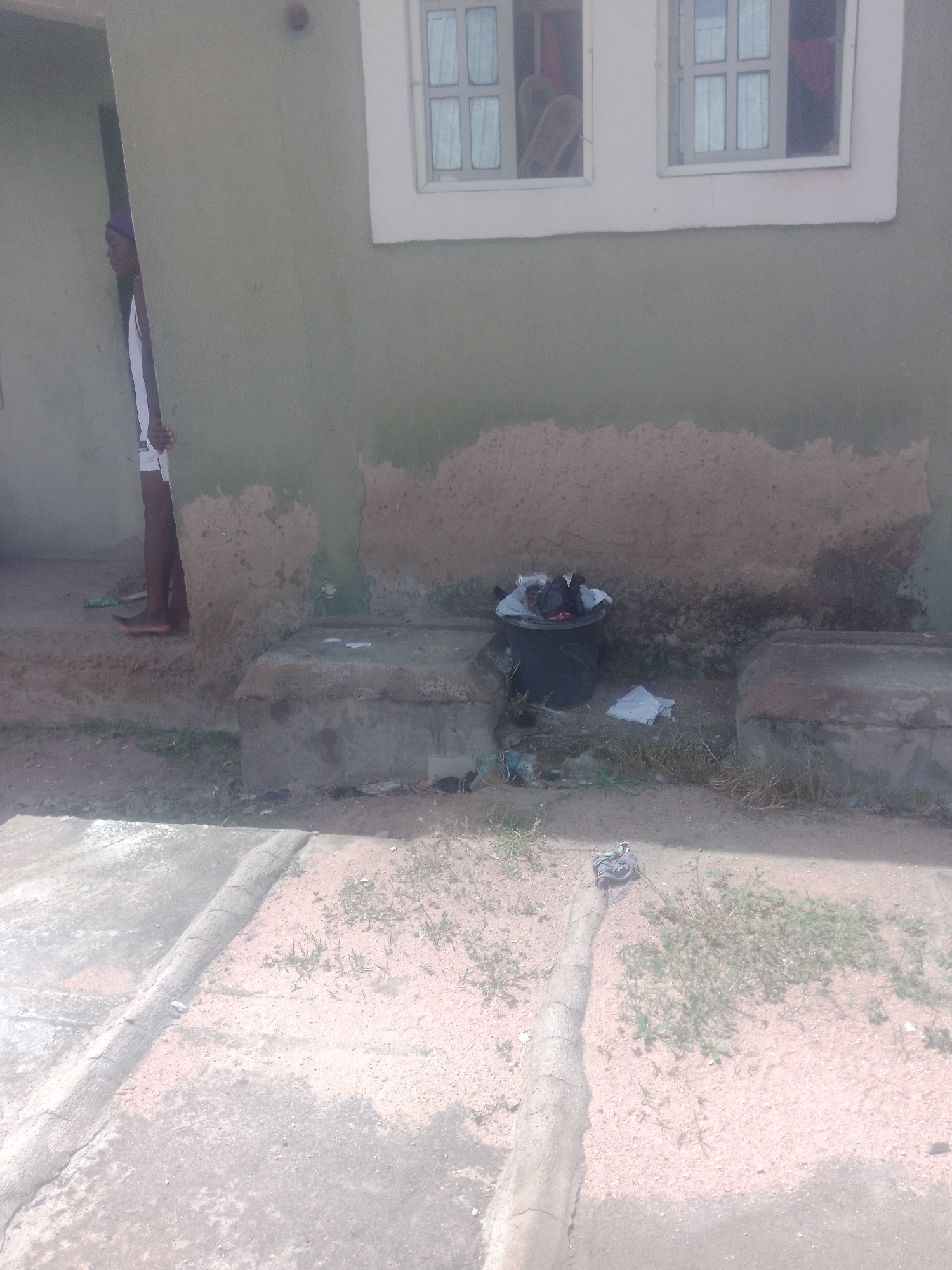 A person is standing in a doorway on the left side of the image, partially obscured by a wall. The person is wearing a white tank top and white shorts. The building in the image is a light green color with a white-framed window above a stained wall. In front of the building, there is a black trash can filled with what appears to be debris. A concrete structure is located to the right of the trash can. The ground in front of the building is a mix of dirt and cracked concrete. There are some plants growing in the sandy areas. The scene appears to be outdoors in a residential area.