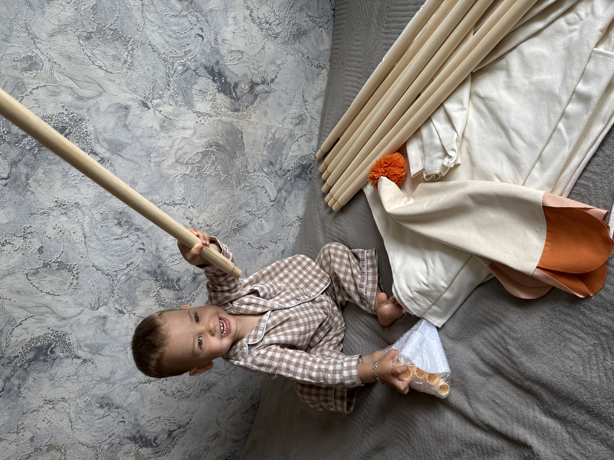 A small child in a brown and white checkered pajama set is seated on a gray blanket next to the canvas and wood frame of a teepee tent. The child is holding a wooden pole in one hand and a bag of wooden dowels in the other. The room's wall is textured and painted in shades of gray. The location appears to be in Rusanovo, Russia.