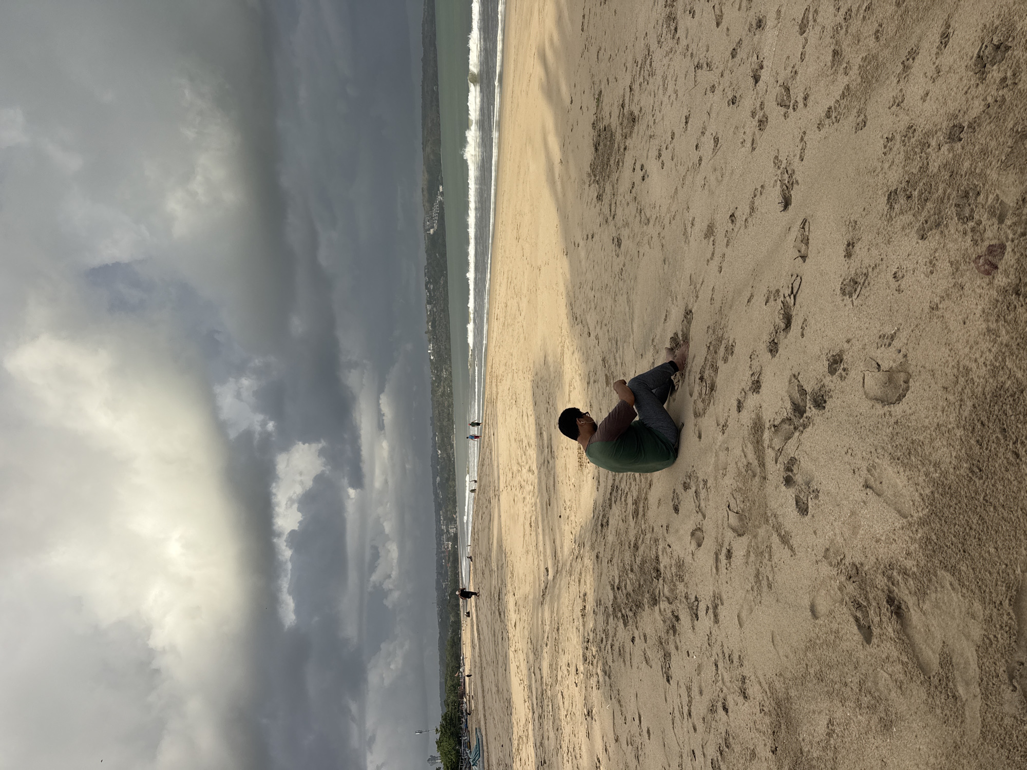 The image depicts a beach scene in Kuta, Indonesia, under a dramatic, overcast sky with patches of bright sunlight. In the foreground, a person is seated on the sand, facing away from the camera and looking down, appearing to be engaged with something in their hands. They are wearing a green and brown t-shirt and grey pants. The sand is a light tan color, marked with numerous footprints and some small indentations.
Further back on the beach, several other individuals are visible, scattered across the sandy expanse. Some are walking, while others appear to be standing or sitting. The ocean stretches out in the mid-ground, with moderate waves breaking onto the shore. The water is a greenish-blue hue.
In the distance, a coastline is discernible, lined with buildings and vegetation, suggesting a developed area beyond the beach. The sky is filled with heavy, dark clouds, interspersed with brighter areas where sunlight is breaking through, casting distinct shadows on the sand. The overall atmosphere is one of natural beauty with a touch of brooding weather.