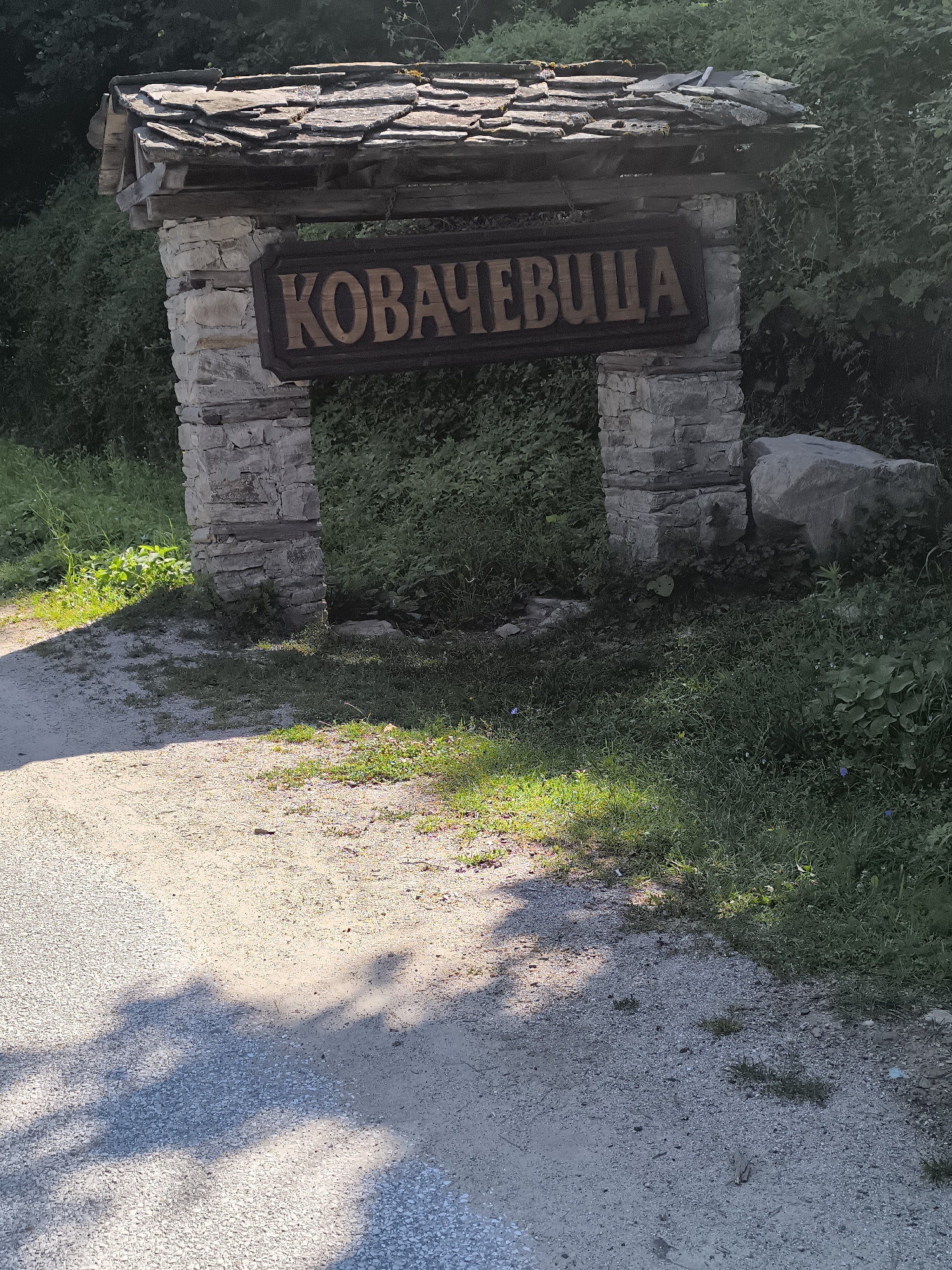 An outdoor shot shows an archway made of rough-hewn stone and wood, with a weathered slate roof. A dark wooden sign with carved letters, reading "КОВАЧЕВИЦА" (Kovachevitsa), is mounted above the archway. The archway is positioned along a dirt path that leads into a wooded area with dense green foliage. Sunlight casts shadows on the path. A large rock is visible to the right of the archway. The overall impression is rustic and natural, suggesting an entrance to a historical or rural location.