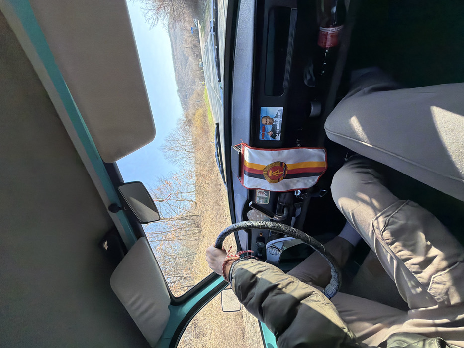 An interior view from a moving vehicle captures a driver and passenger traveling on a paved road in Dobromirka, Bulgaria, under a clear blue sky. The driver, wearing a dark olive jacket and light brown trousers, has their left hand on the steering wheel, which is covered with a black textured grip. A red and black braided bracelet is visible on the driver's left wrist. The passenger's legs, also in light brown trousers, are partially visible on the right.
The vehicle's dashboard is dark. A small flag featuring the East German national emblem (hammer, compass, and wreath of rye) against the black, red, and gold stripes of the GDR flag is suspended from the rearview mirror mount. To the right of the flag, a rectangular object, possibly a mobile phone, rests on the dashboard. Adjacent to this, a sticker or small sign displays an image of Vladimir Putin giving a thumbs-up, accompanied by Russian text reading "Xaxaxa! Ты не работаешь!" ("Hahaha! You don't work!"). A red bottle lies horizontally in a storage compartment to the far right of the dashboard. The sun visors are retracted upwards. The interior window frames are a distinct teal color.
Outside the vehicle, a two-lane asphalt road stretches forward, curving slightly. The road is bordered by dry, earthy terrain and hills covered with mostly bare deciduous trees, indicative of late autumn or winter. Distant vehicles are visible on the road ahead. The horizon is defined by undulating hills under a bright, cloudless sky.