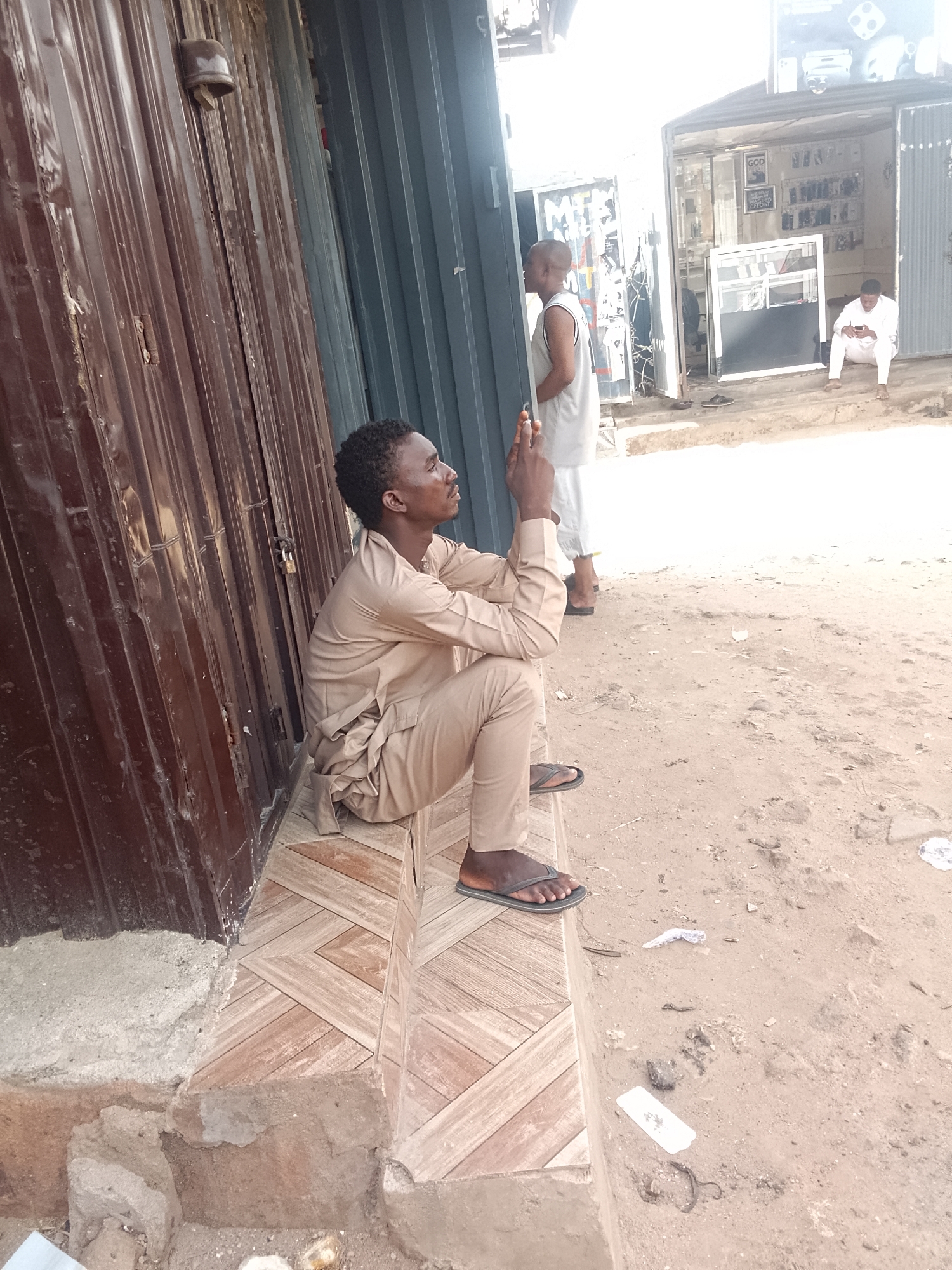 The image depicts an outdoor scene in what appears to be Gombe, Nigeria. A man wearing a light brown long-sleeved shirt and trousers is sitting on a tiled step outside a corrugated metal structure. He is looking upwards with his hands clasped together. Another man in a white tank top and shorts is standing near the structure. In the background, there is an open-front shop with a person sitting inside. Debris is scattered on the ground.