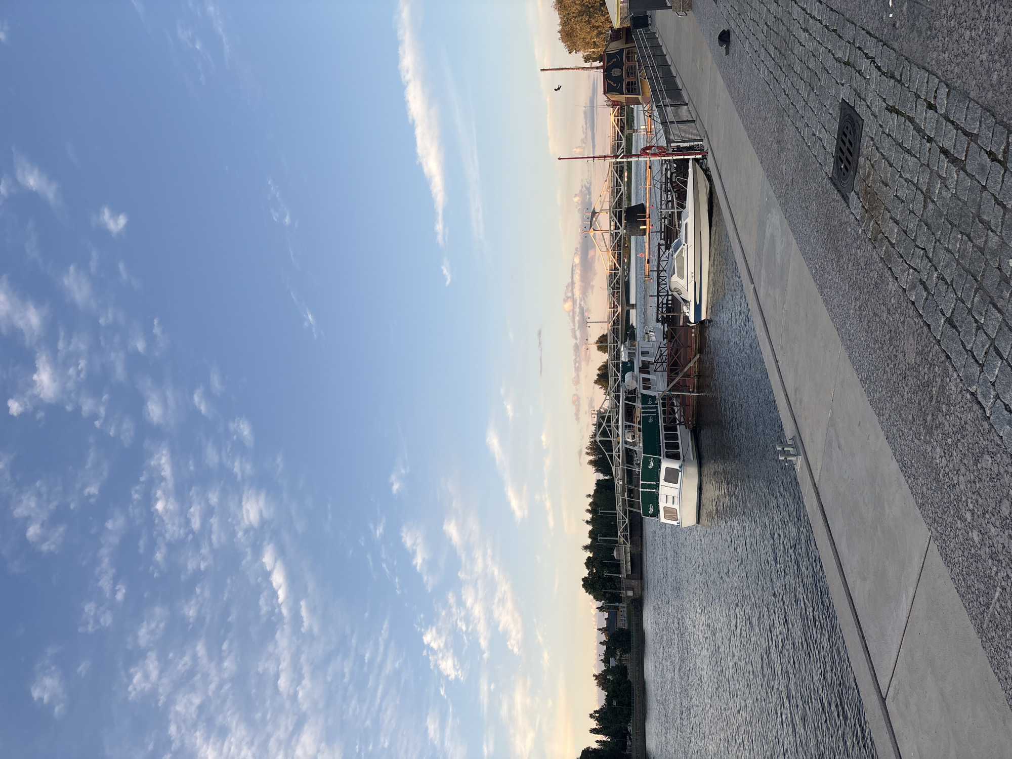 This is a vertical, eye-level shot of a harbor in Pori, Finland, on a partly cloudy day with a bright blue sky. The water is calm, and several boats are docked along the embankment. In the foreground, a concrete walkway transitions to a cobblestone path. A drain grate is visible on the cobblestones. A black cap lies on the cobblestones to the right of the drain.
The main focus is a large white passenger boat with green accents docked on the left side of the image. A smaller white speed boat is also docked in front of the passenger boat. Further in the background, a bridge spans the water, with trees lining the opposite shore. Another boat, designed to look like a pirate ship, is docked on the right side of the harbor. A bird is flying in the sky above the pirate ship. The sun is low in the sky, casting a warm glow on the clouds and creating soft shadows. The scene appears peaceful and serene.