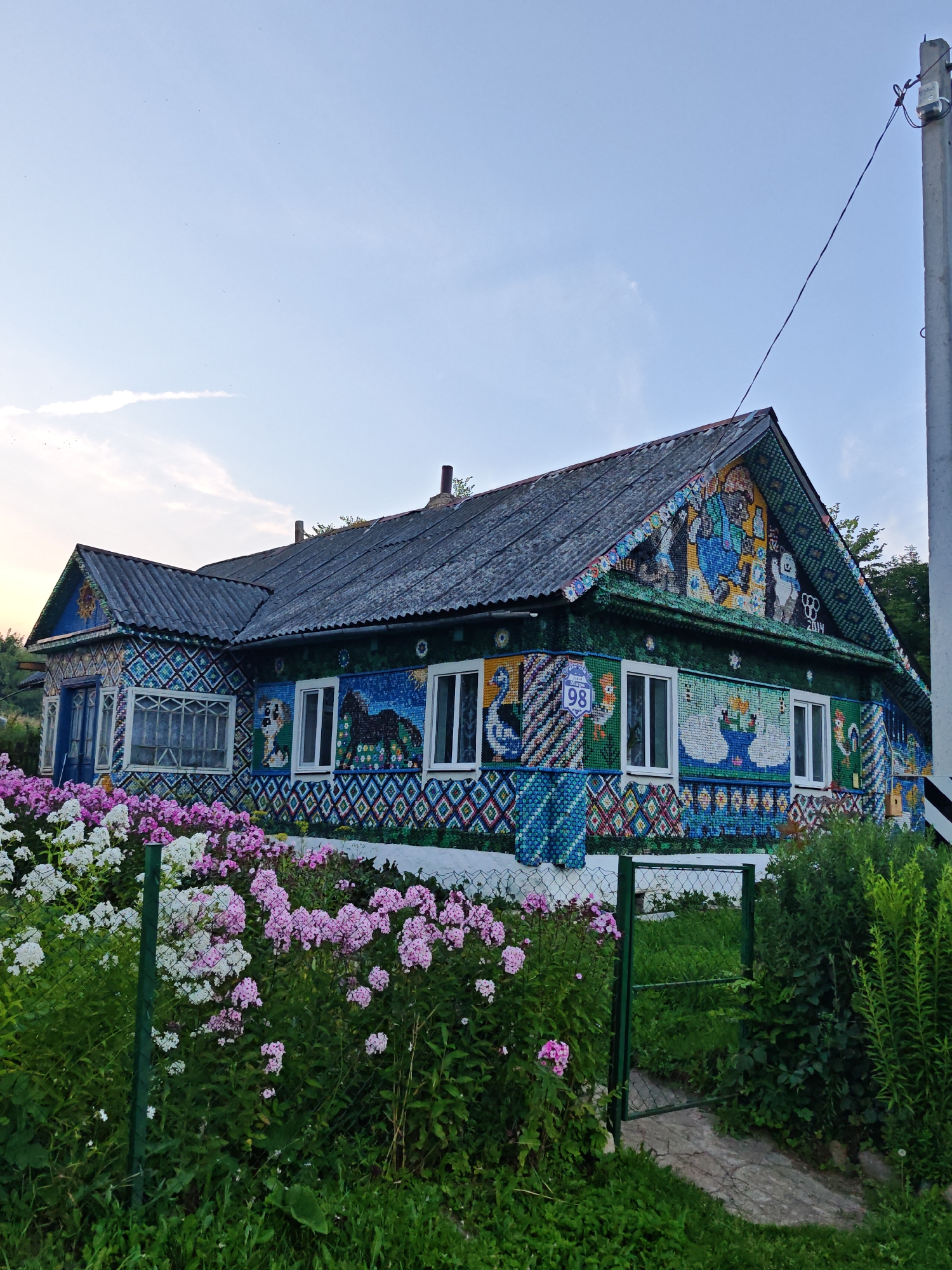 The image captures a brightly decorated house in Valozhyn Rural Council, Belarus. The house's exterior walls and roof are adorned with a mosaic made of bottle caps, forming various colorful patterns and images, including animals like a horse and swans, as well as human figures. The house has several windows, and a blue door is visible on the left side. In front of the house, a lush garden is blooming with pink and white flowers, separated from the house by a green metal fence. A dirt path leads to a green gate, beyond which more greenery and trees are visible. A utility pole with wires stands to the right of the house, and the sky above is a clear blue with a few wisps of clouds. The scene appears to be set in a rural or semi-rural area.