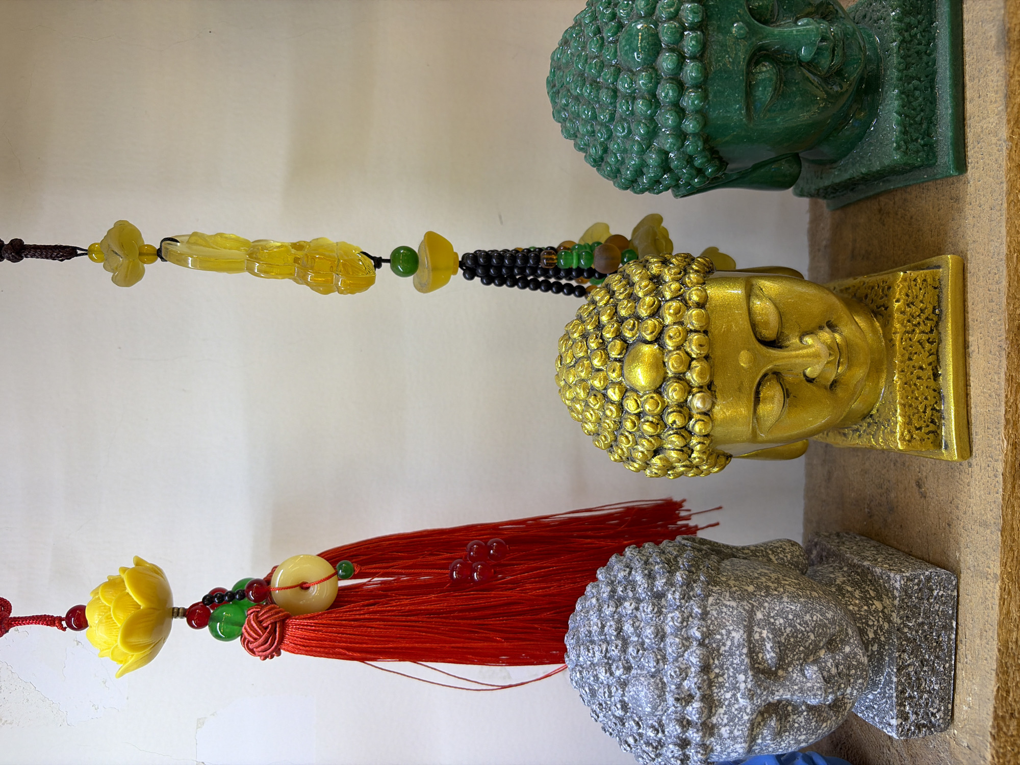 Three Buddha head busts are displayed on a wooden shelf. The bust on the left is a speckled gray stone. The center bust is metallic gold with intricate curls for hair. The bust on the right is a translucent green. Above the busts, to the left, hangs a red tassel ornament with a yellow lotus flower and other decorative beads. To the right of that, a yellow beaded ornament with a yellow carved flower and dark beads hangs. The background is a plain, textured off-white wall. The scene appears to be indoors, likely a shop or decorative display. This image was taken in Da Nang, Vietnam.