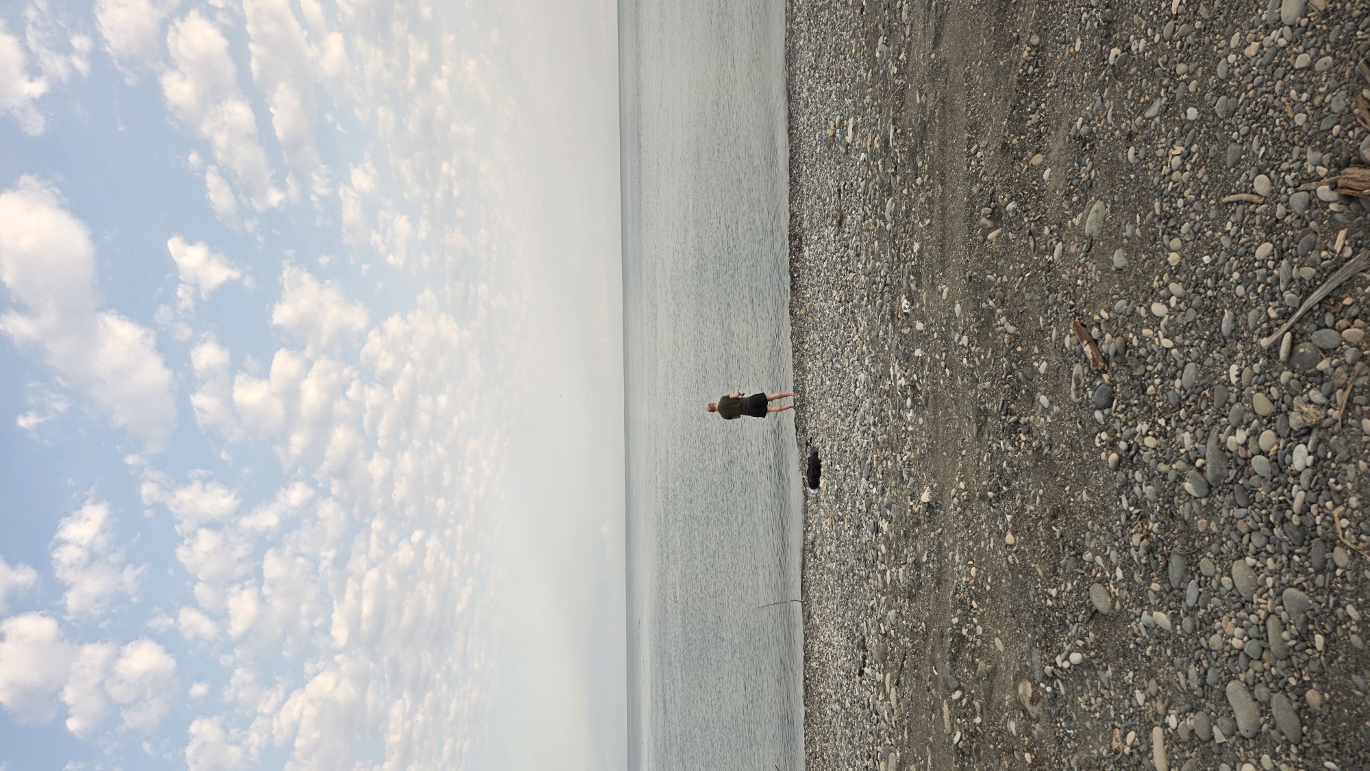 A lone person stands on a pebbly beach at the edge of a calm, grey body of water. The person, wearing a dark t-shirt and dark shorts, faces away from the camera, towards the water, and appears to be holding a fishing rod. The sky above is a pale blue, dotted with scattered, fluffy white clouds. The beach is covered in a dense layer of small, smooth grey and white pebbles, with a few scattered pieces of driftwood. A black, possibly deflated tire is visible on the beach near the water's edge. The scene suggests a quiet, natural environment. The location is a beach in Batumi, Georgia, which is characterized by its pebble beaches and views of the Black Sea.