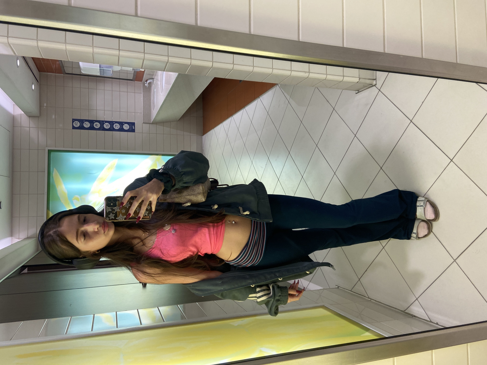 A young woman with long brown hair and wearing a pink crop top, dark pants, and open-toe sandals is taking a selfie in a restroom mirror. She is holding a smartphone with a patterned case in her right hand. She is wearing headphones and a jacket. The restroom features white tiled walls, a hand-washing station, and signage in both English and Arabic indicating it is likely in Dubai, United Arab Emirates. The floor is also tiled with a diamond pattern.