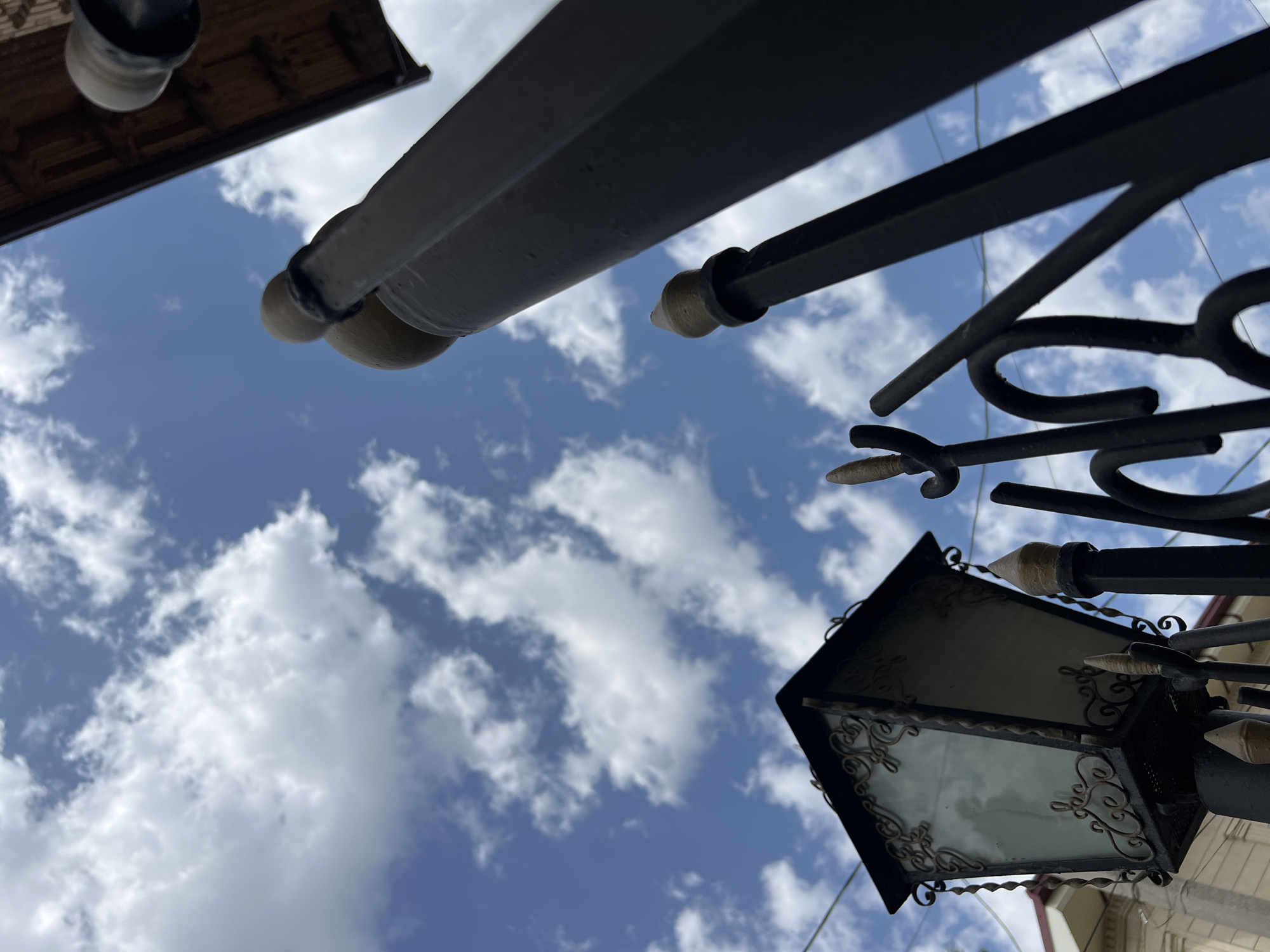 The image, taken from a low angle, shows a cloudy blue sky with a decorative, black wrought-iron fence and lamppost in the foreground. The lamppost has an ornate, hexagonal lantern with frosted glass panels. Above and to the right, a dark wooden overhang is visible. The scene suggests an outdoor urban setting, possibly a residential area or a public space with architectural details. The overall impression is of a bright, daytime scene with a focus on the contrast between the man-made structures and the natural sky.
