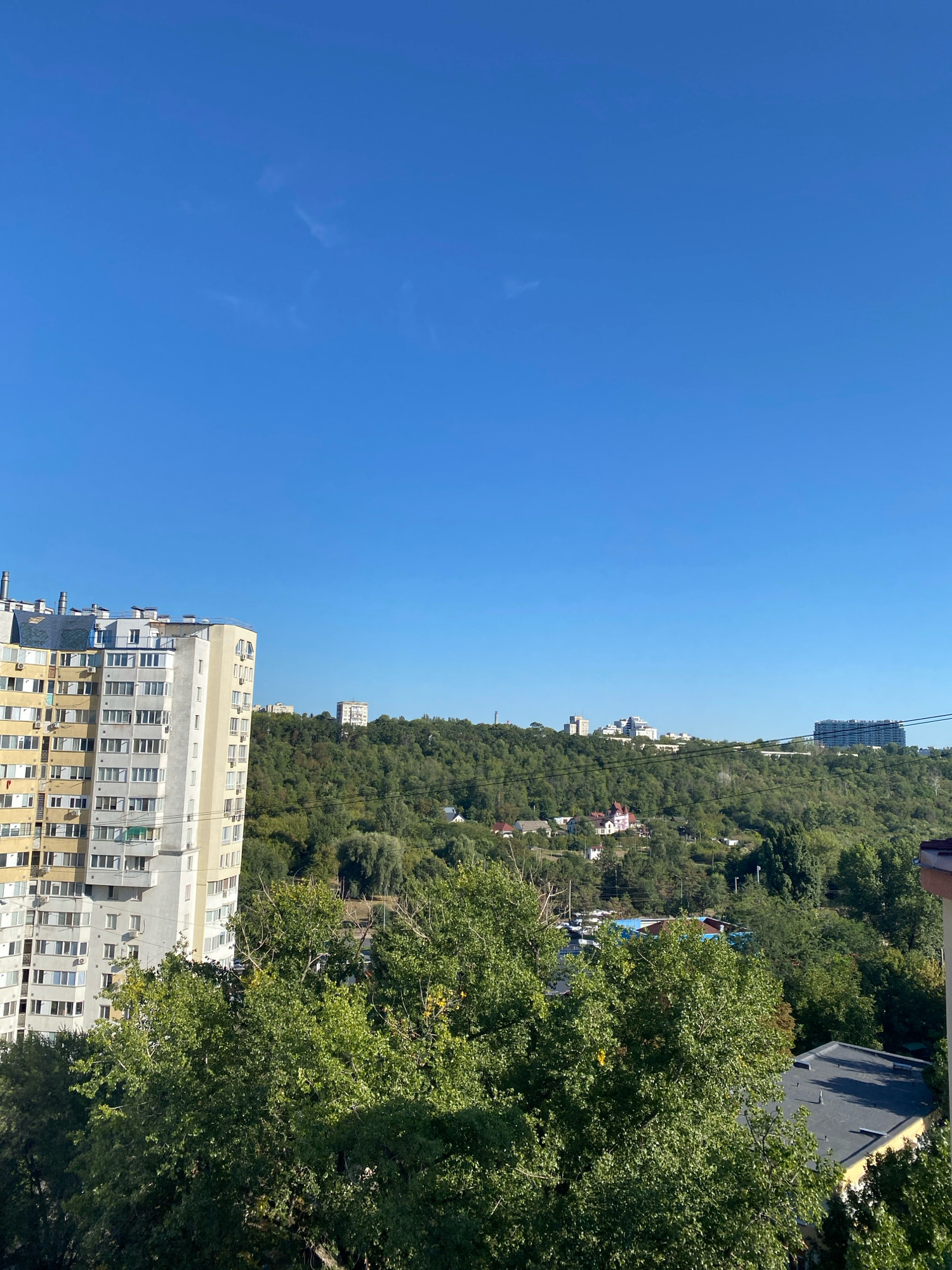 This is a landscape photograph taken from a high vantage point, likely a balcony or window, looking out over a residential area in Chișinău, Moldova. The sky is a clear, bright blue with a few wispy clouds.
In the foreground, on the left side of the image, is a tall, multi-story apartment building. It has a light yellow and beige facade with numerous windows and balconies. Air conditioning units are visible on some of the balconies. The building appears to be an older Soviet-era design, common in many Eastern European cities.
Beyond the apartment building, a dense, green forested hillside rises. Scattered amongst the trees, especially on the lower slopes, are smaller houses and some more modern buildings visible further up on the ridge. The foliage is lush and appears to be in full summer or early autumn bloom.
On the far right of the image, the corner of another building is visible, with a dark, angled roof. This building is closer to the viewer than the main apartment block.
The overall impression is one of a city with significant green spaces and a mix of older and newer architecture. The image captures a sunny day with good visibility, offering a panoramic view of the urban and natural landscape.